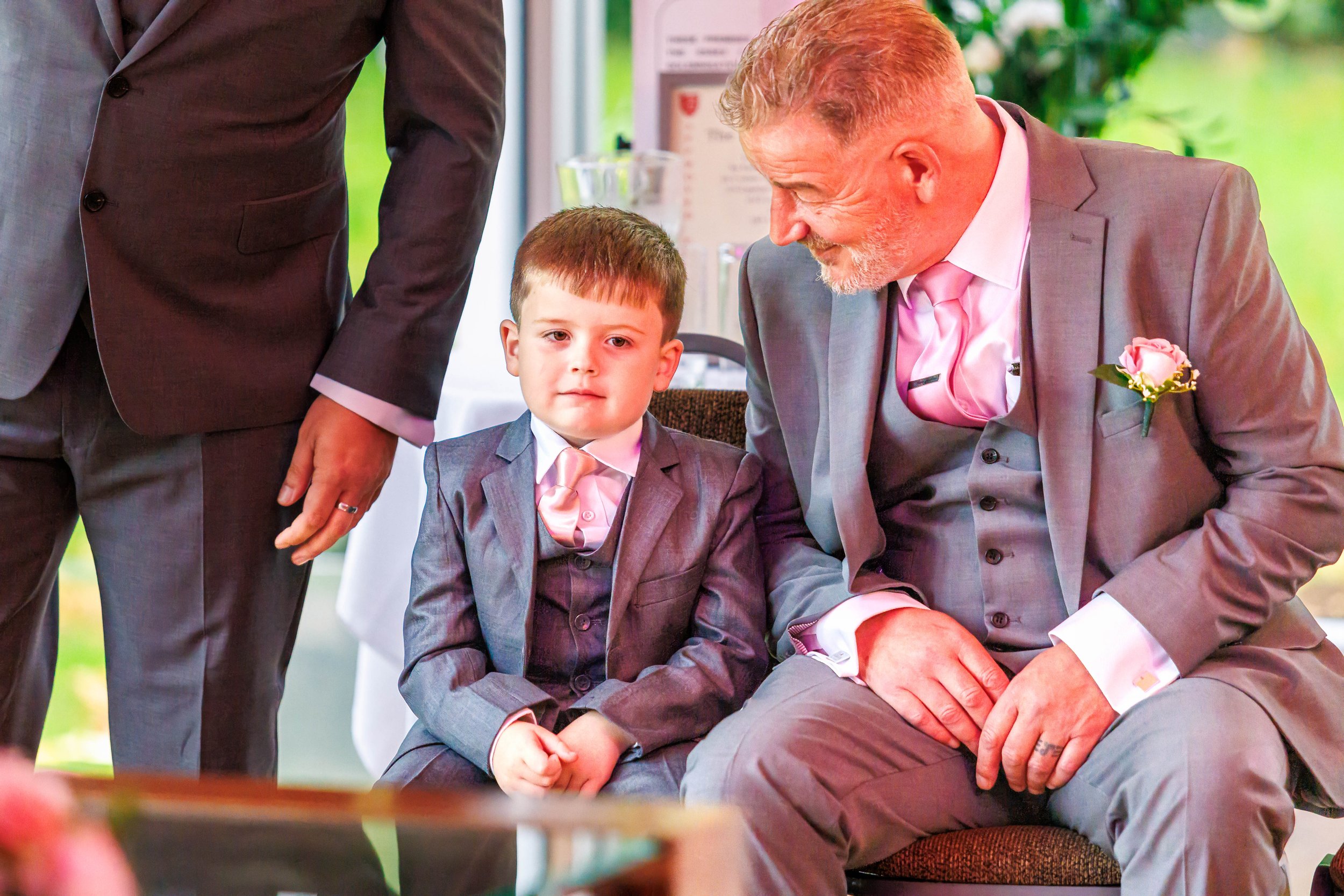 A young boy and an older man sitting in gray suits with pink ties, at a formal event.