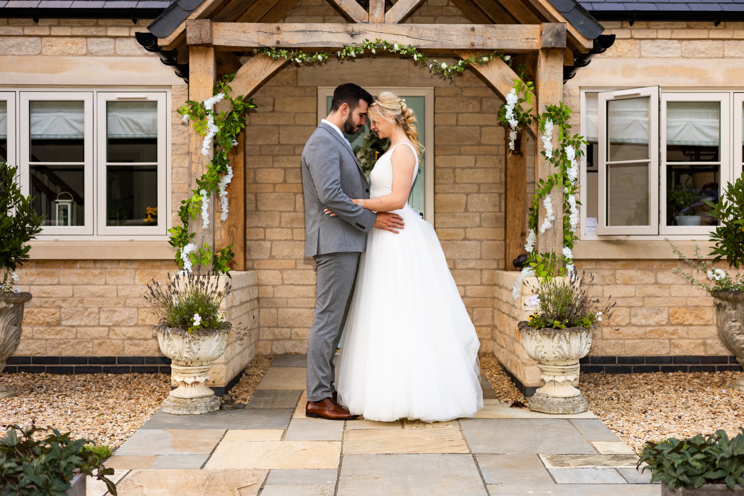Bride and groom embracing under a wooden arch decorated with green and white flowers outside a stone building.
