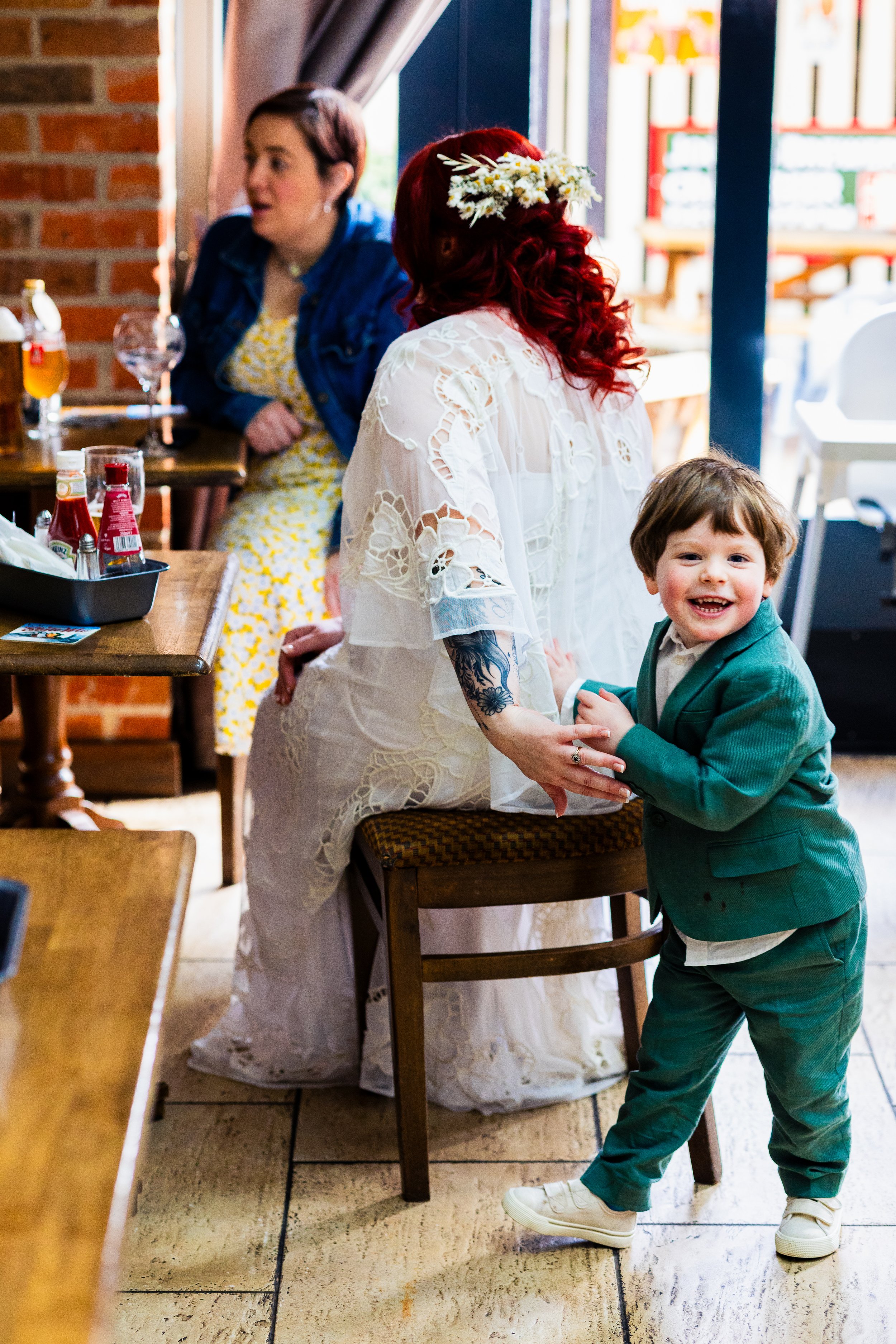 A child wearing a green suit smiles and holds the hand of a person with red hair seated indoors, while another person sits at a nearby table in a casual setting.