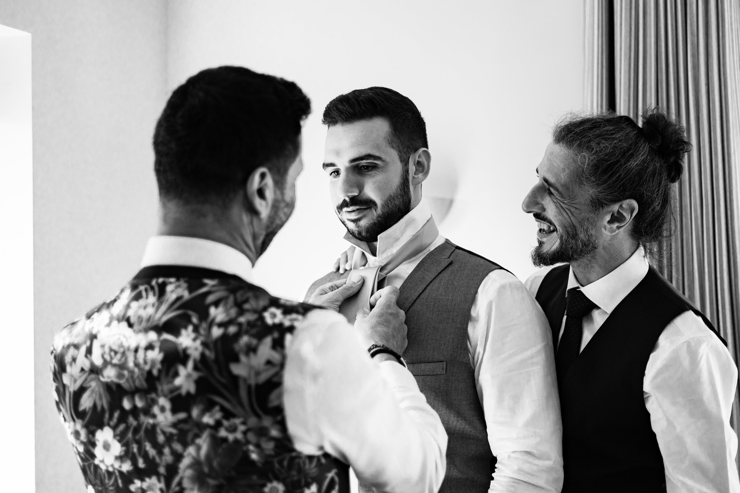 Three groomsmen in formal attire, with one adjusting a tie for another, smiling and preparing for an event. A fatgher adjusting his son.