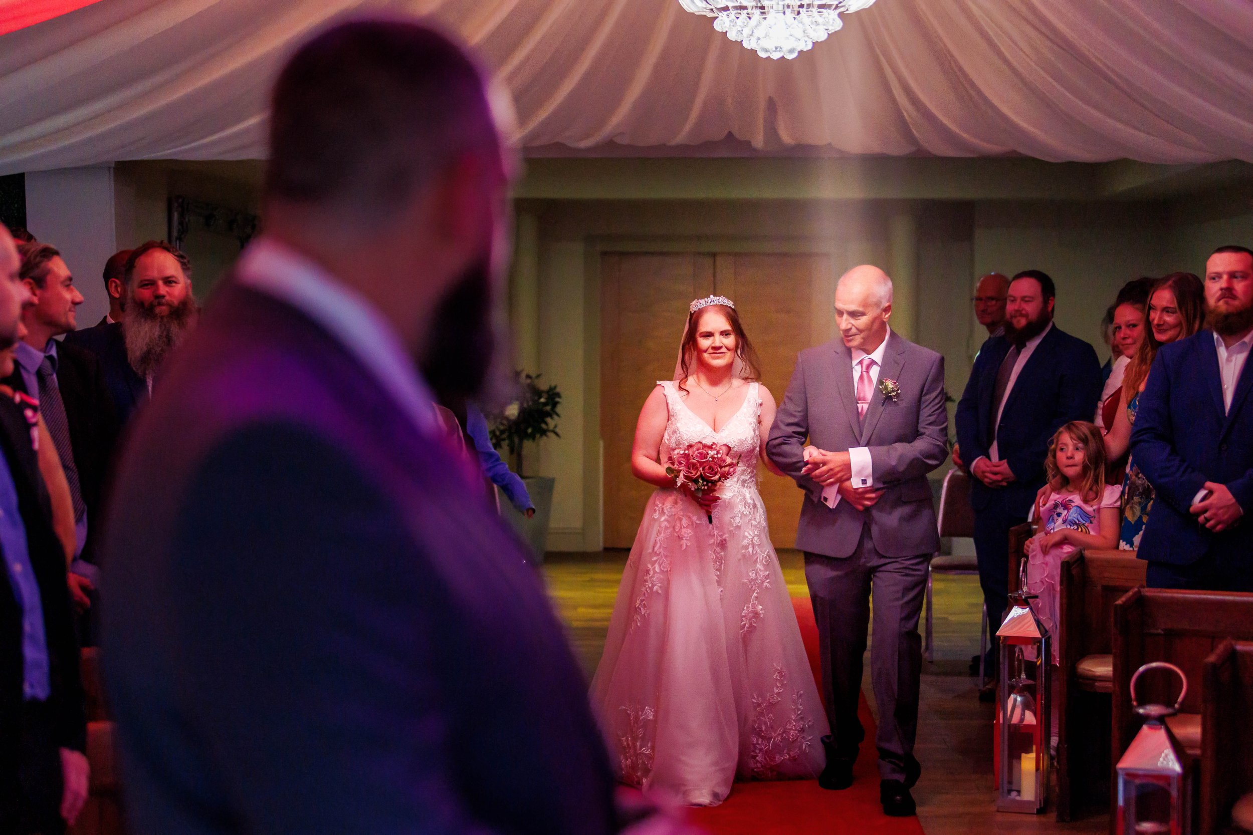 A bride in a white dress is walking down the aisle with an older man, likely her father. She's holding a bouquet of flowers. They are surrounded by guests in formal attire inside a venue with draped fabric on the ceiling and a chandelier. A man in fo