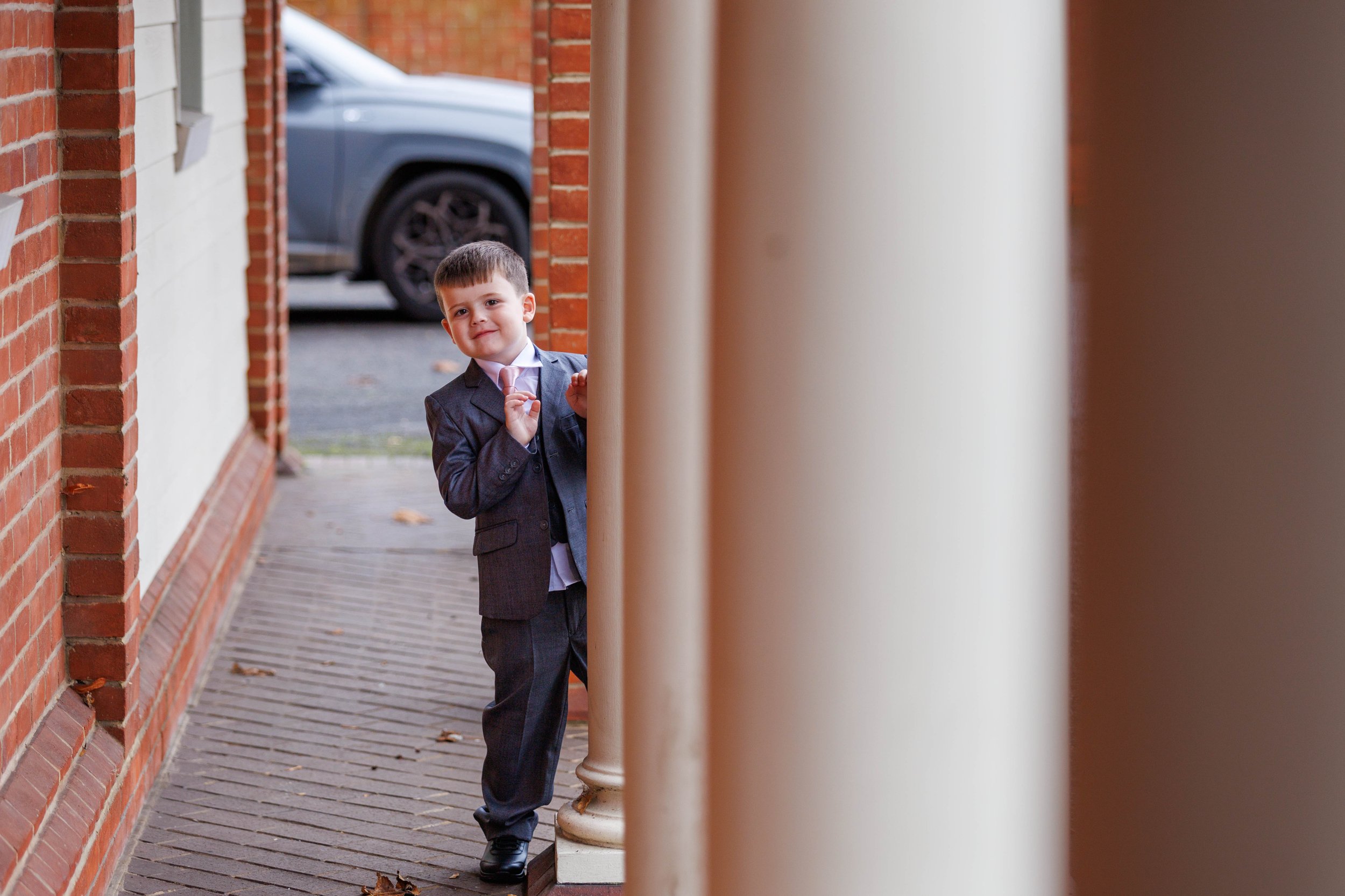 Young boy in a suit holding a pink bow tie, standing by a brick building and columns, with a gray car in the background.