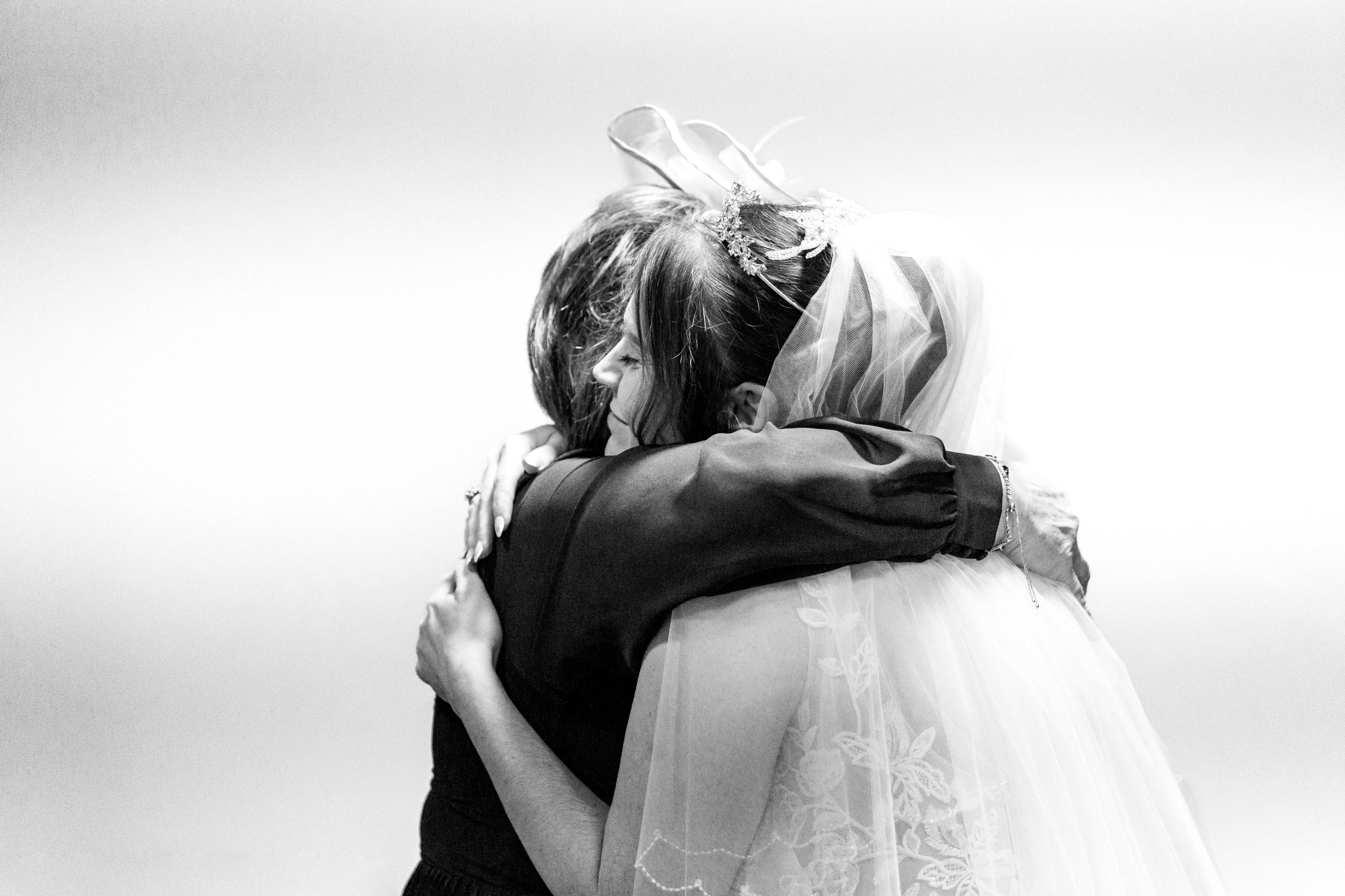 A black and white image of a bride embracing another person. The bride is wearing a veil and ornate headpiece.