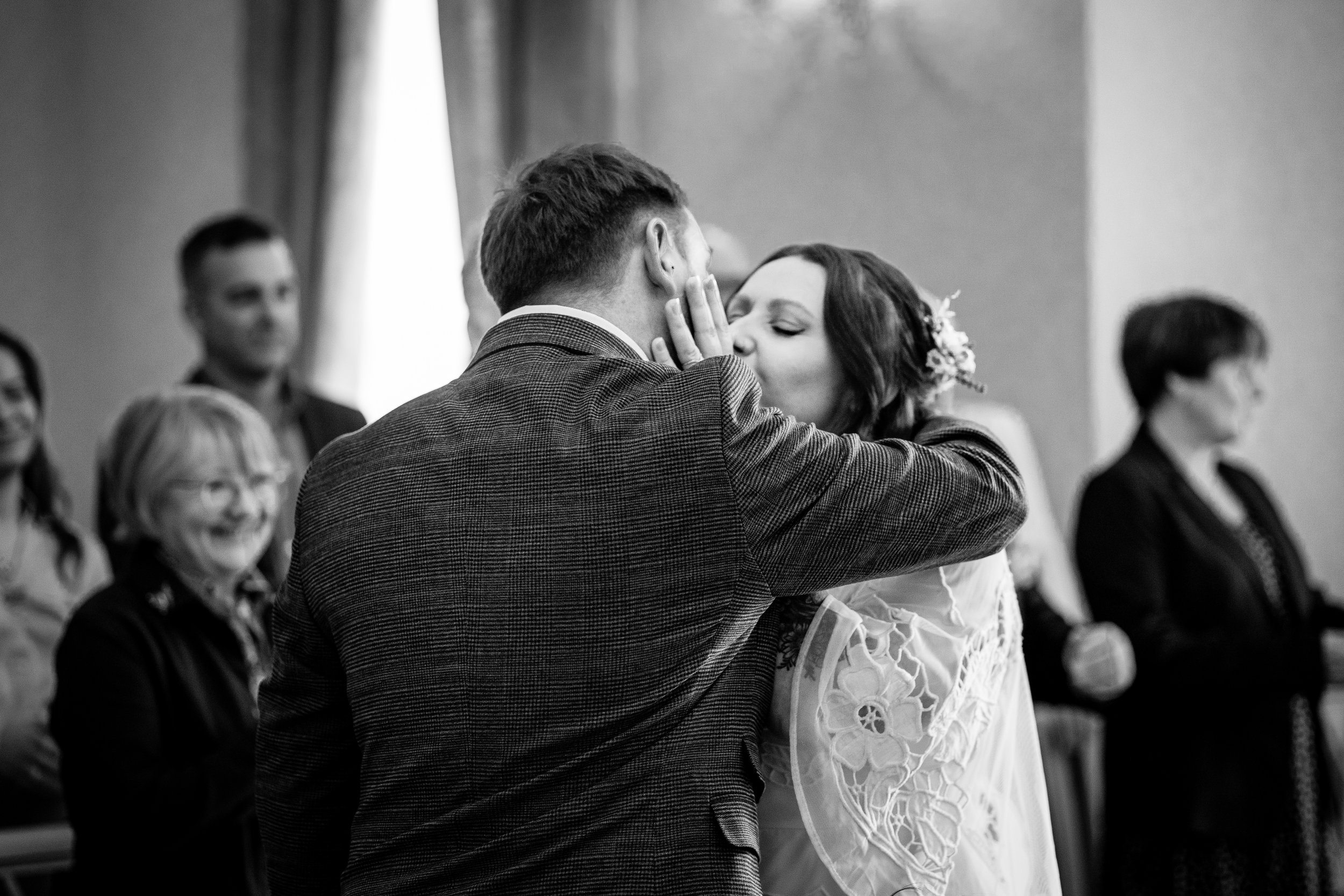 A bride and groom kissing at a wedding ceremony with guests smiling in the background, black and white image.