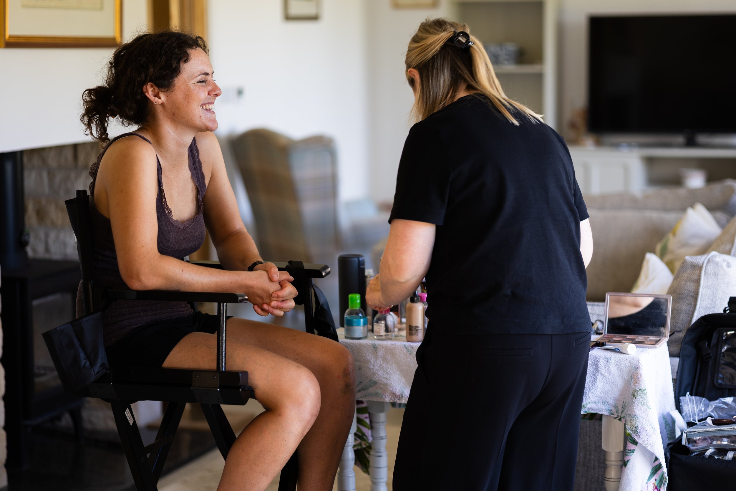 Woman sitting in a chair smiling while a makeup artist applies products in a living room setting.
