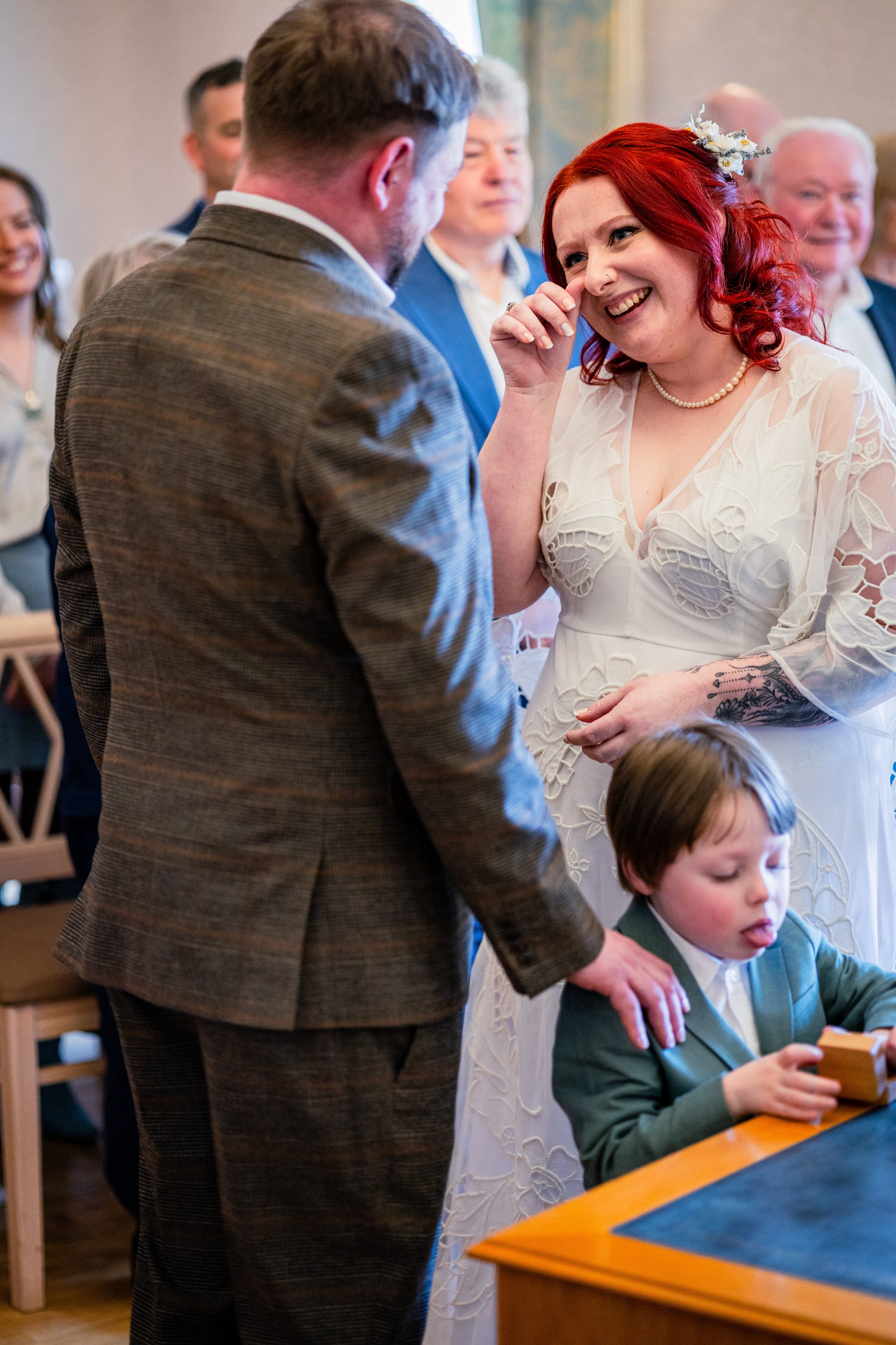 Bride smiling while wiping tear, groom in suit facing her, child playing with box, and guests in background at a wedding ceremony.