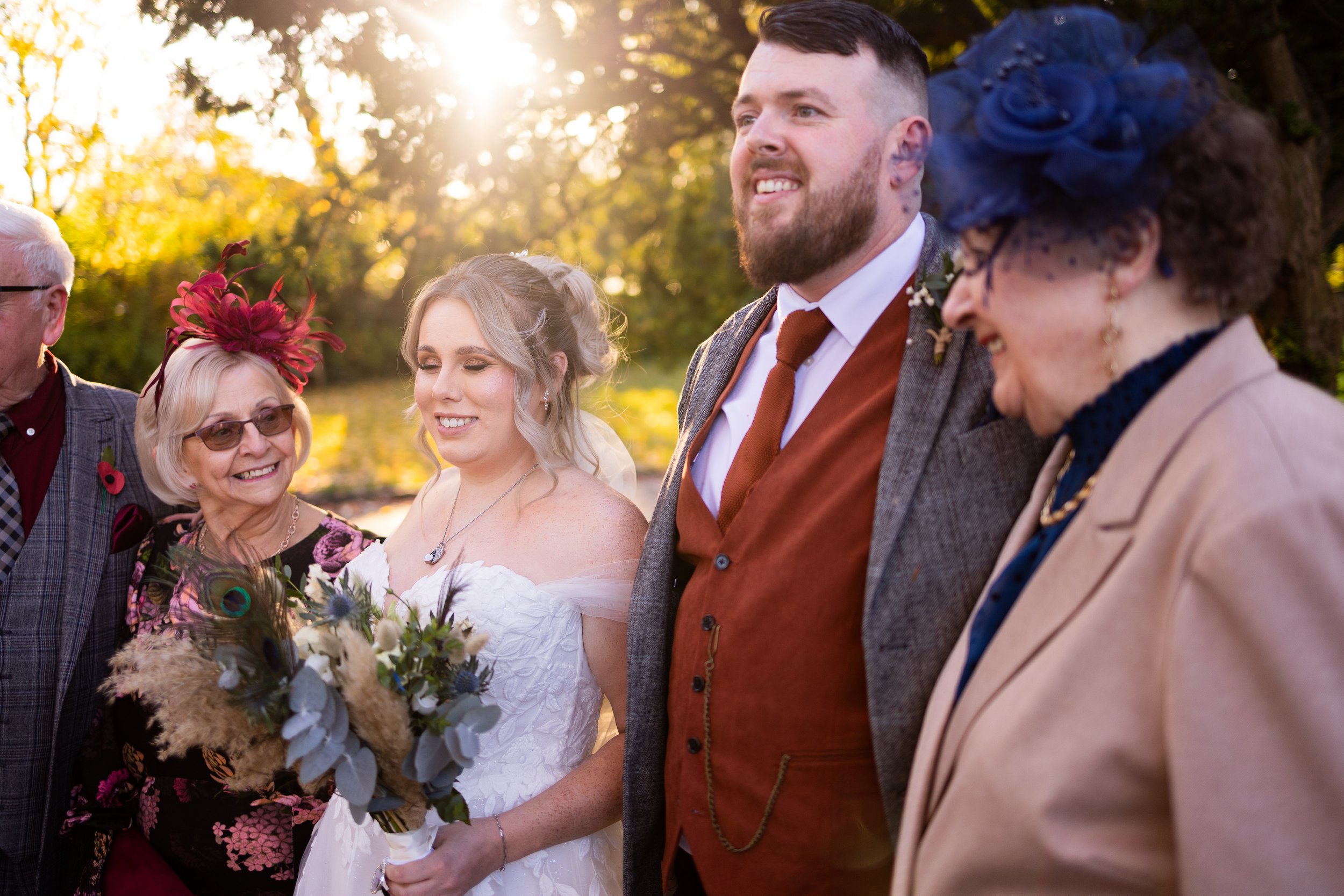 A bride and groom standing outdoors with family members, smiling in a sunlit setting. The bride holds a bouquet of flowers, while the groom wears a brown vest and tie. Two older women beside them wear colorful hats. The atmosphere is joyful and celeb