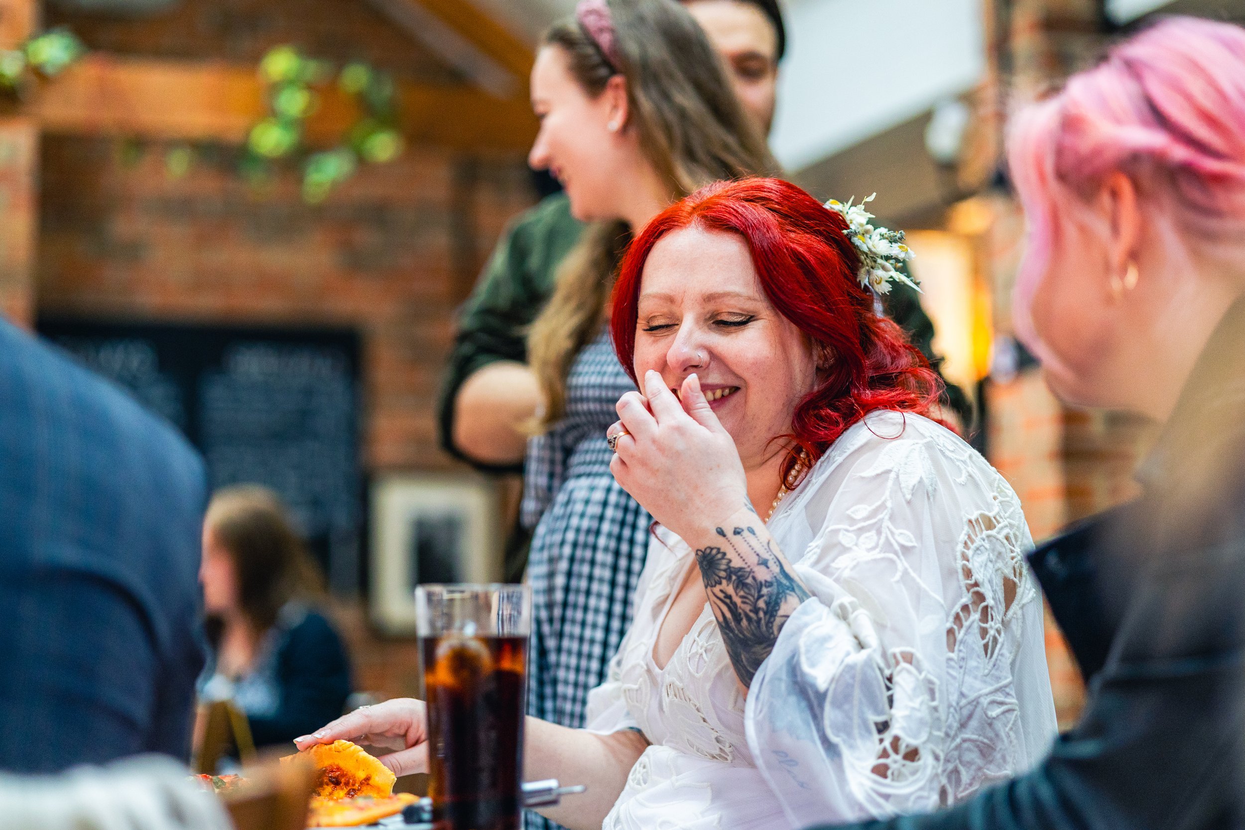 Smiling woman with red hair in a white dress, sitting at a table with a drink and food, interacting with others in a lively setting.