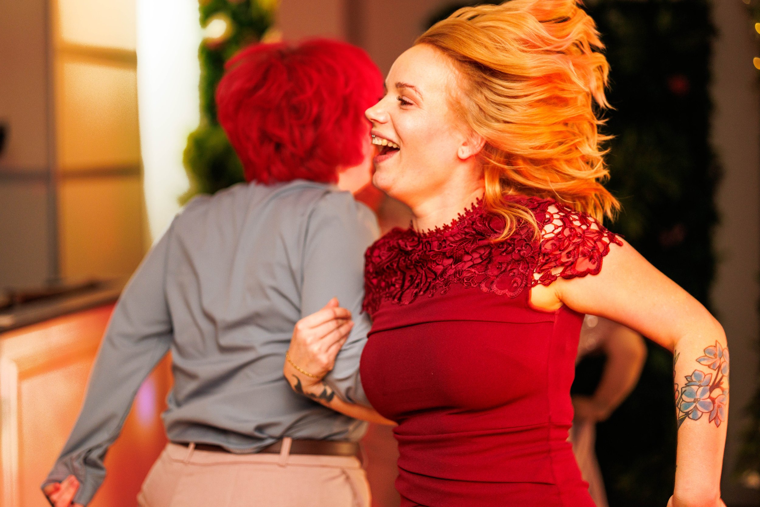 Two women dancing energetically, with one wearing a red dress and the other in a blue shirt, in a lively indoor setting.