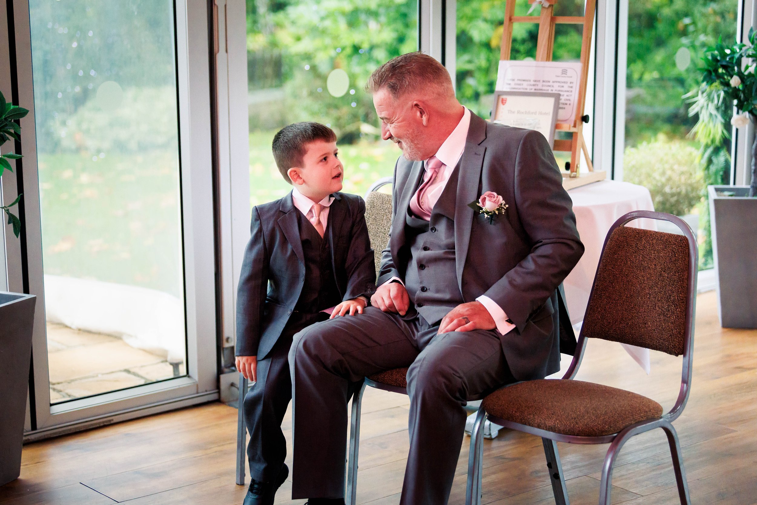A young boy and a man in matching gray suits with pink ties sit next to each other in a bright room, looking at each other affectionately. The man has a boutonniere on his lapel. There's a large window with a view of greenery behind them.