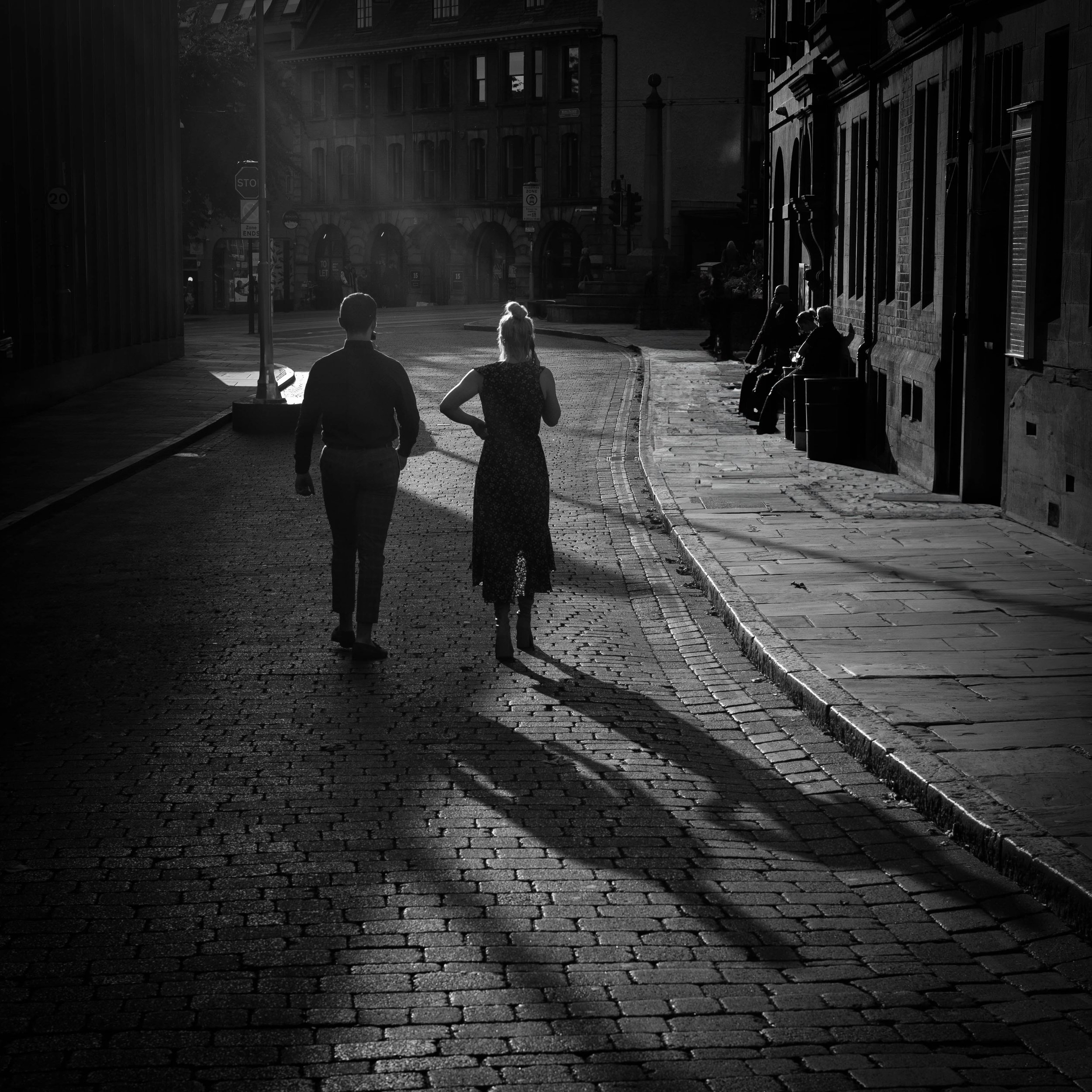 Silhouetted couple walking on a cobblestone street at sunset, casting long shadows, with historic buildings and pedestrians on the sidewalk.