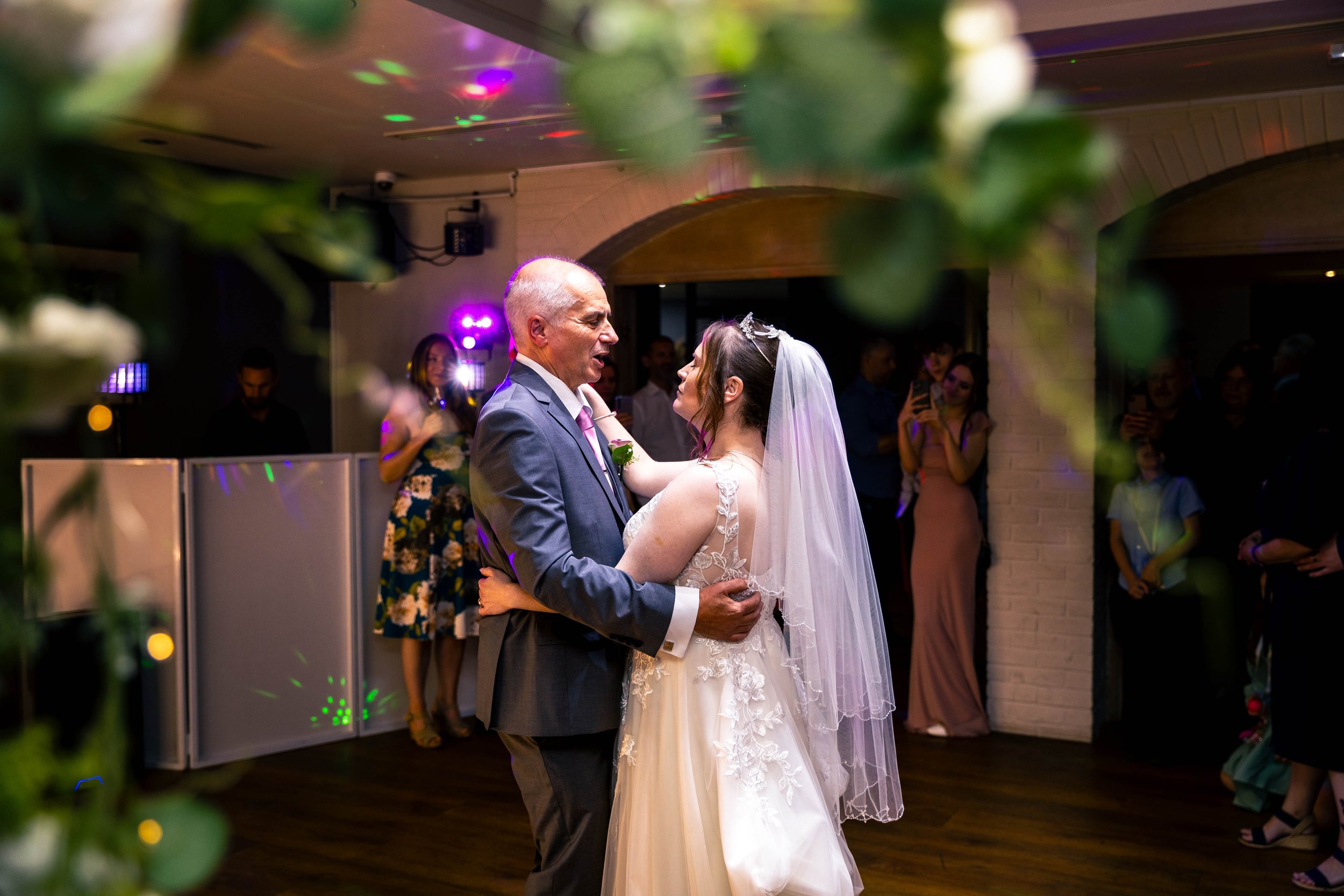 A bride and an older man dancing at a wedding reception, surrounded by guests and soft colorful lighting.