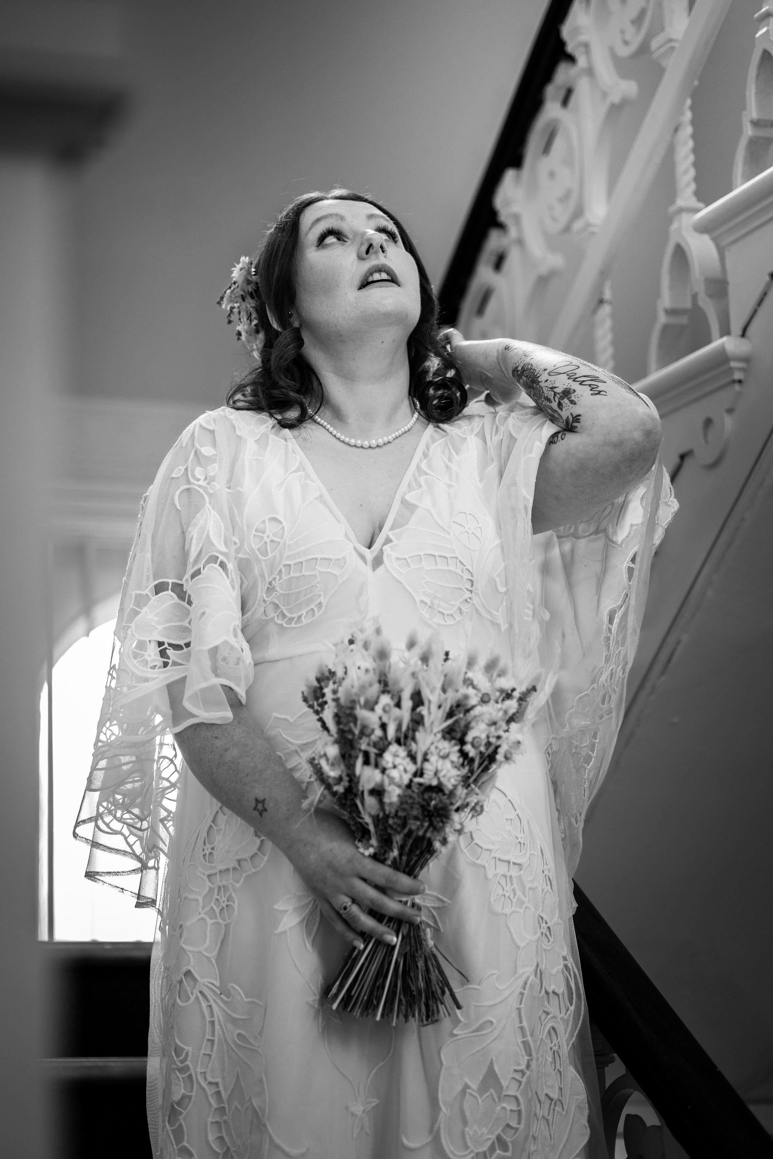 Bride in lace gown holding bouquet on staircase, black and white photo.