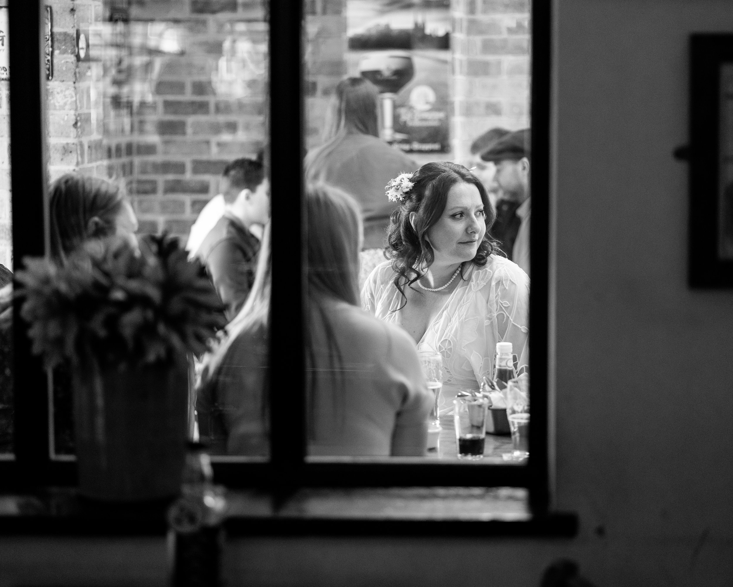 Black and white photo showing a woman with flowers in her hair sitting at a table indoors, viewed through a window. Several other people are visible, engaged in conversation or dining. The setting has a rustic brick interior.