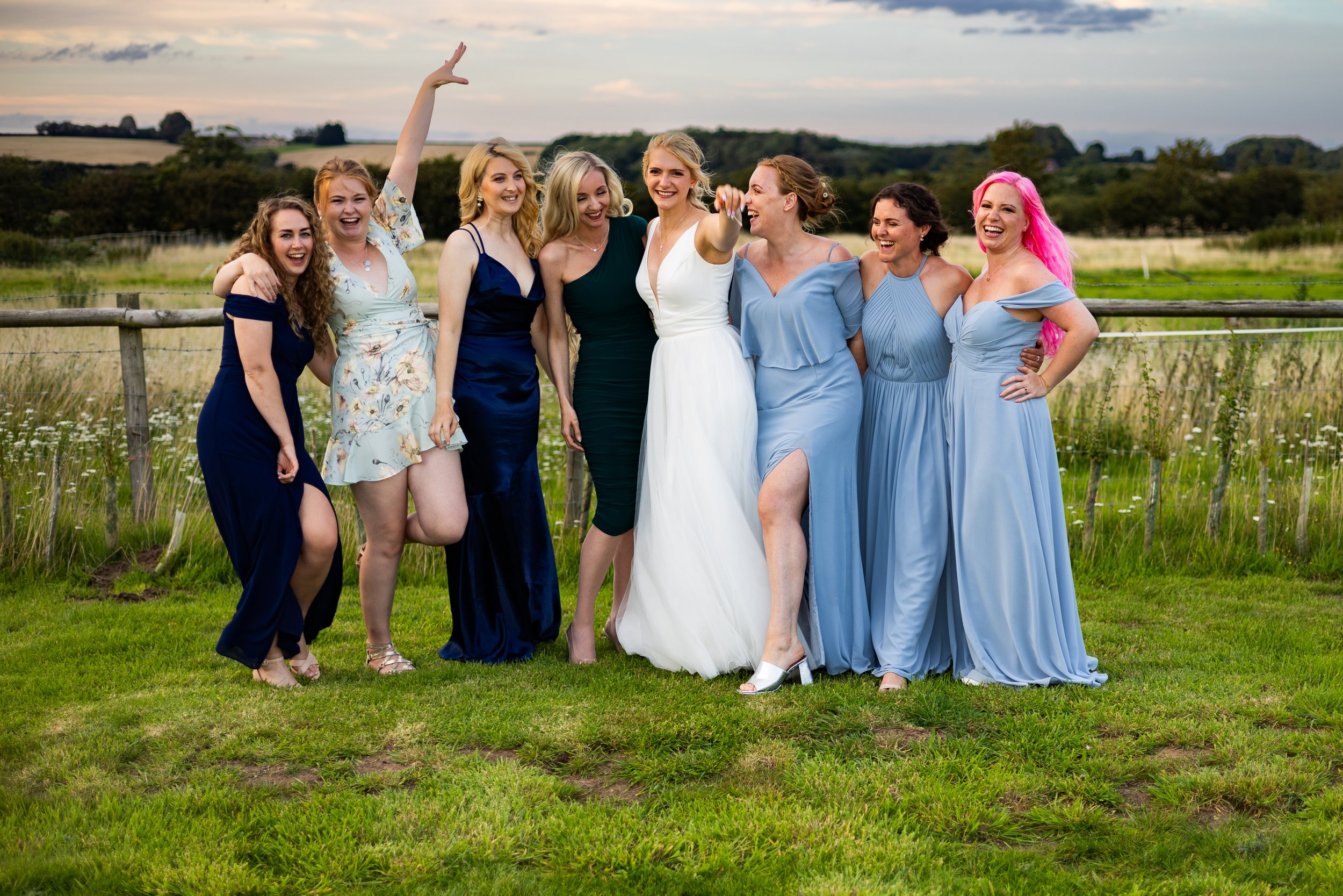 Group of eight women posing outdoors in formal dresses, with a rural landscape in the background.