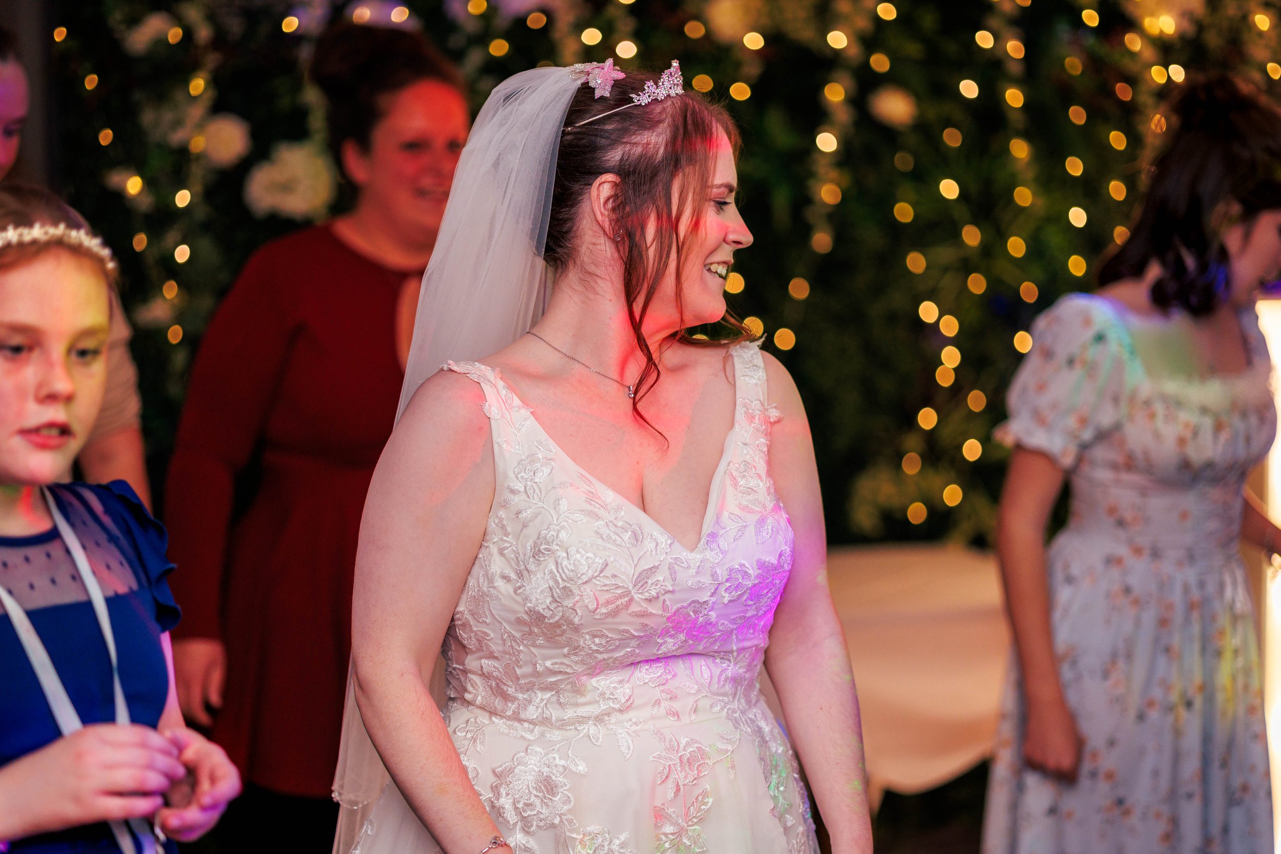 A bride in a white lace wedding dress with a veil smiles while surrounded by guests at a celebration. The background features decorative lights and floral elements.