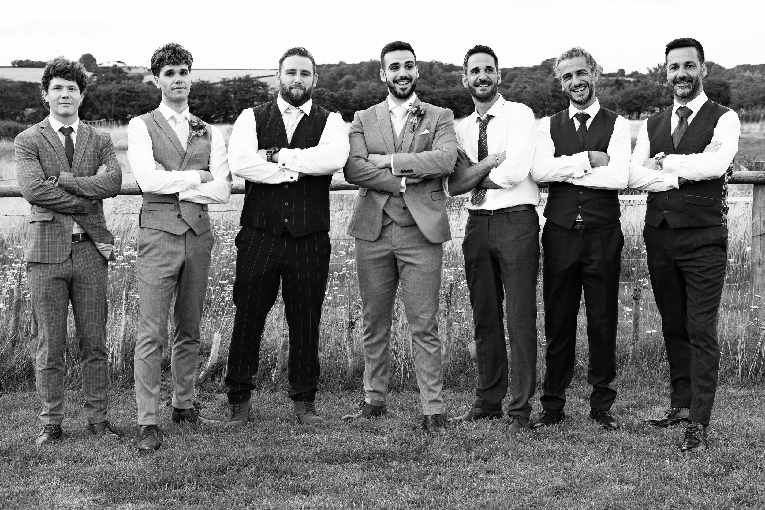 Black and white photo of seven men in formal attire, standing in a row with arms crossed in a grassy outdoor setting.