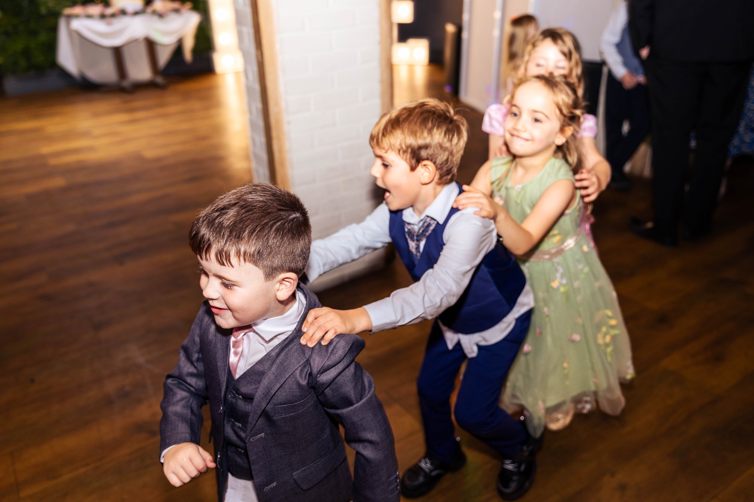 Children in formal attire playing a conga line at a party with wooden floor and decorated background.