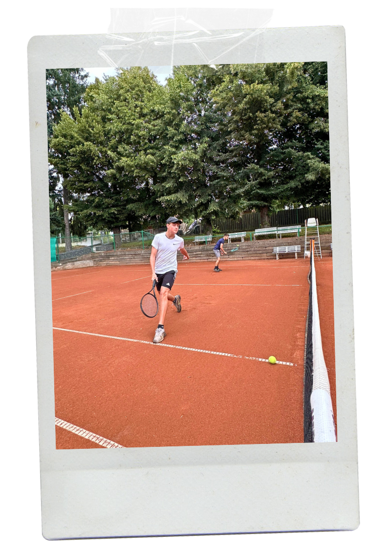 Two boys playing tennis on an orange clay court surrounded by trees, with benches in the background.