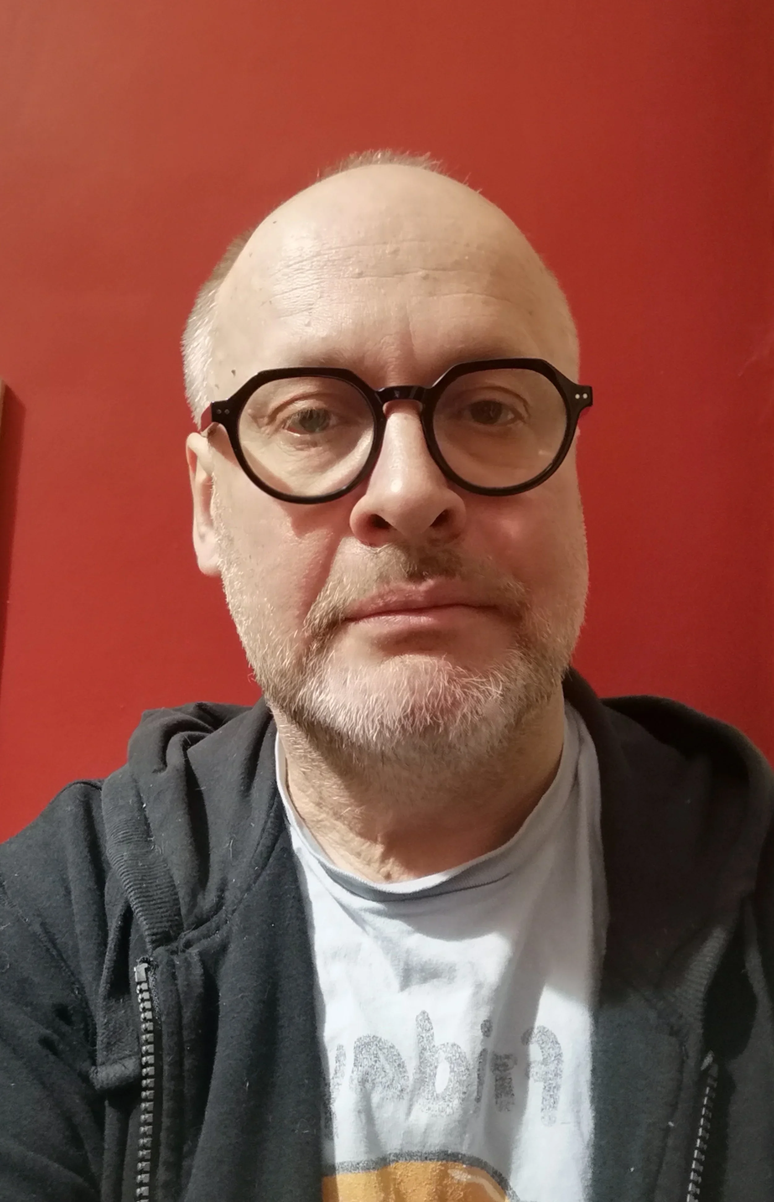 A middle-aged man with glasses, a beard, and short hair, wearing a black hoodie and a white t-shirt, taking a selfie against a red wall.
