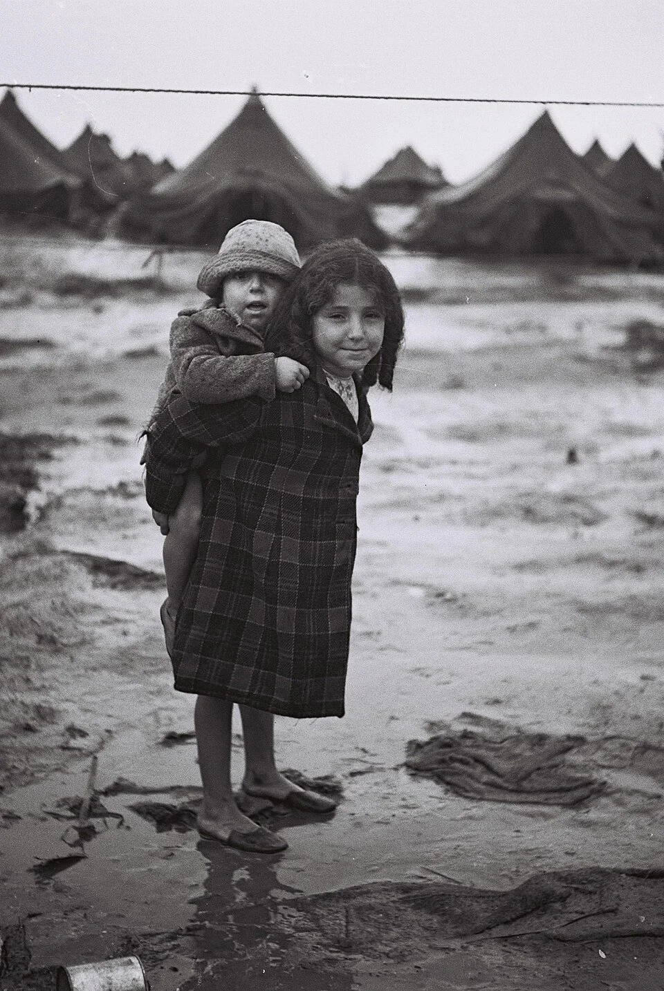 A YOUNG GIRL CARRYING HER BROTHER WALKING THROUGH THE MUD AT THE BEIT LID CAMP. BEIT LID CAMP, ISRAEL עברית:  ילדה נושאת על גבה את אחיה הצעיר ביום חורפי במחנה העולים בבית ליד.