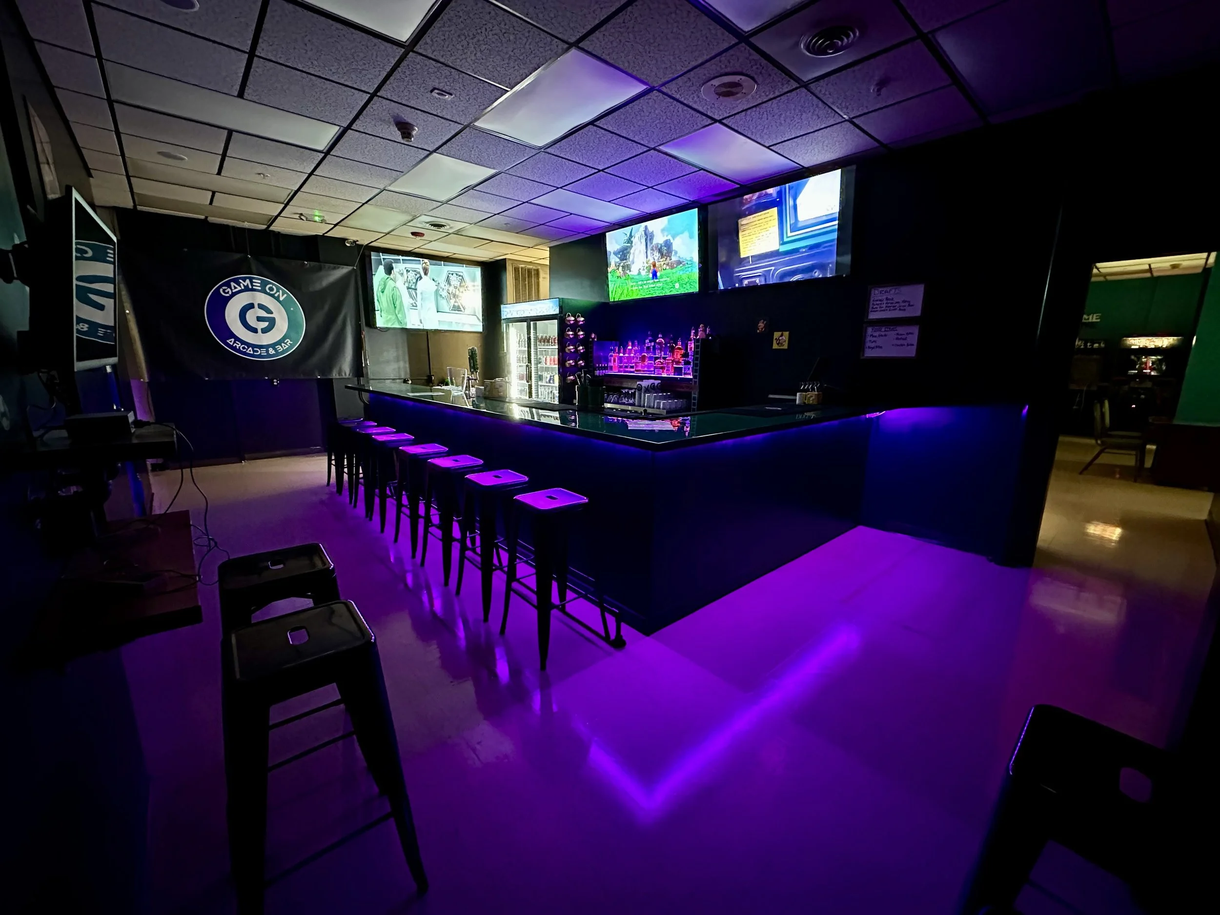 A dimly lit bar with a curved counter illuminated by purple lights, with multiple TV screens displaying video game scenes mounted on the wall behind the bar, surrounded by dark walls and ceiling.