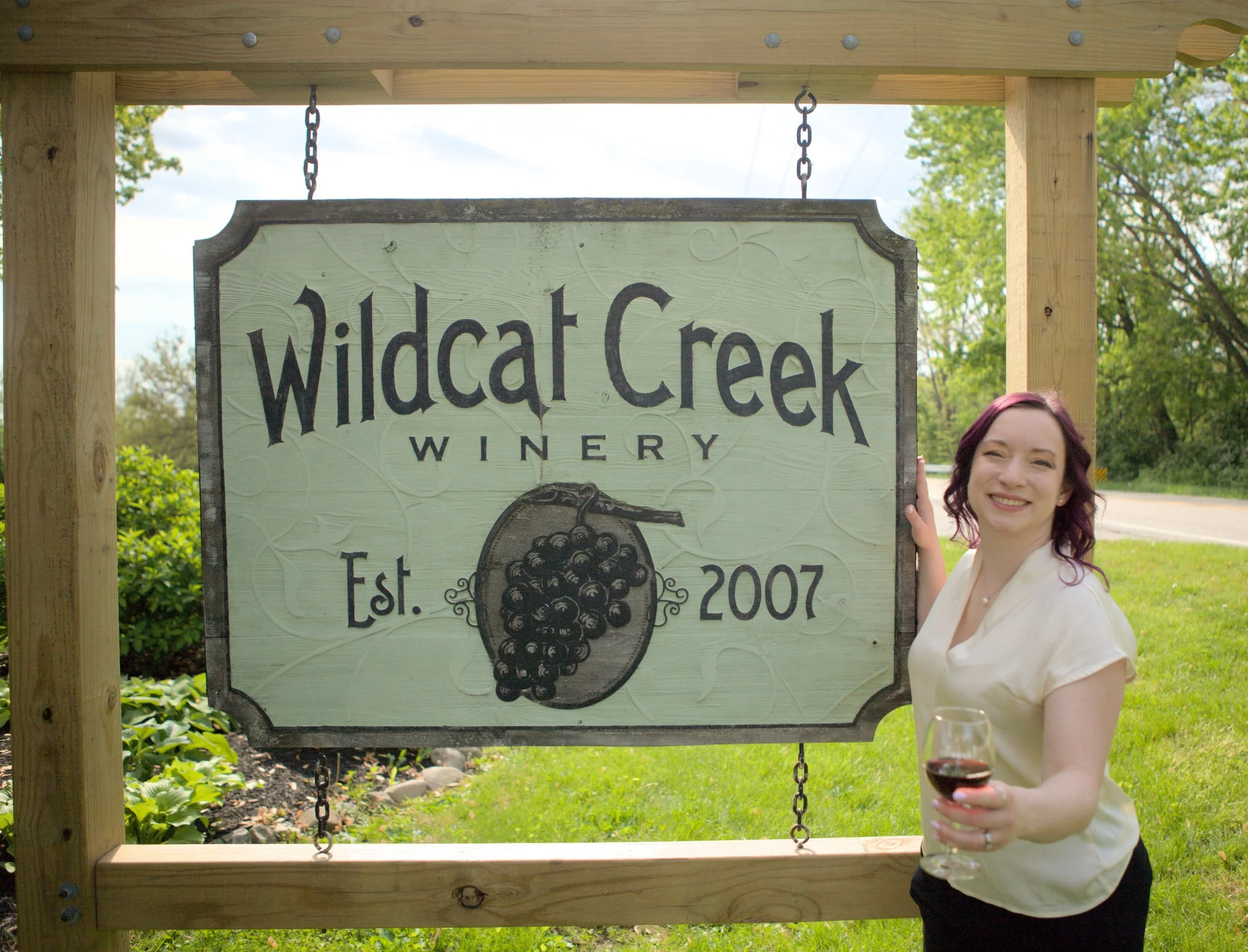 Sign for Wildcat Creek Winery hanging from a wooden structure, with a winemaker smiling and holding a glass of red wine as she stands outdoors near green trees. Lafayette Indiana. Home of Purdue University.