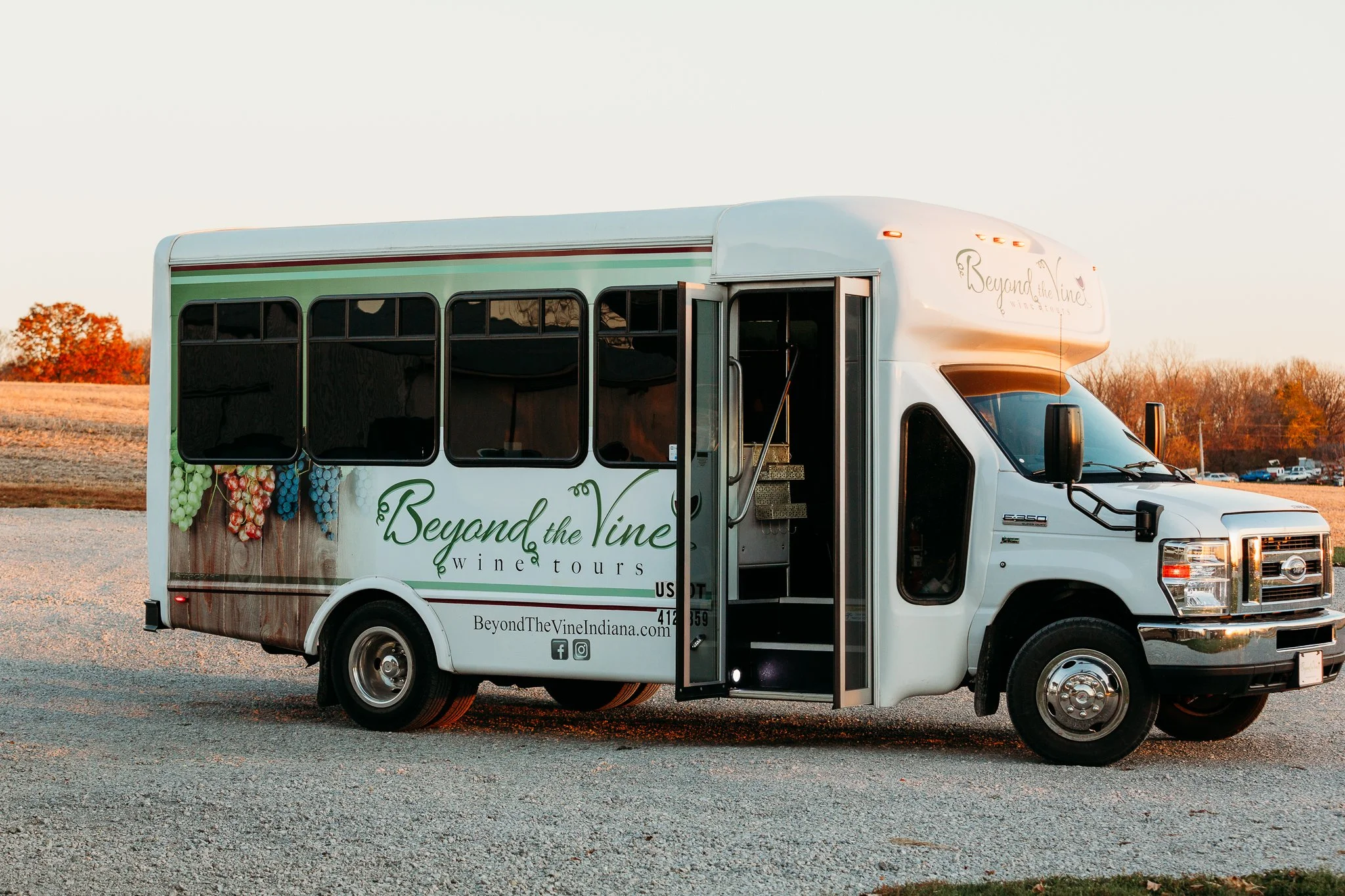 A white luxury shuttle bus with the logo and text "Beyond the Vine Wine Tours" parked outdoors during sunset, with a door open revealing steps inside. 14 passenger transportation. Luxury wine tours.