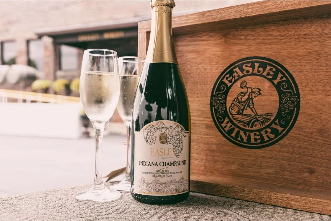 A bottle of Easley Indiana Champagne and two filled champagne glasses on a table outdoors, with a wooden Easley Winery box in the background.
