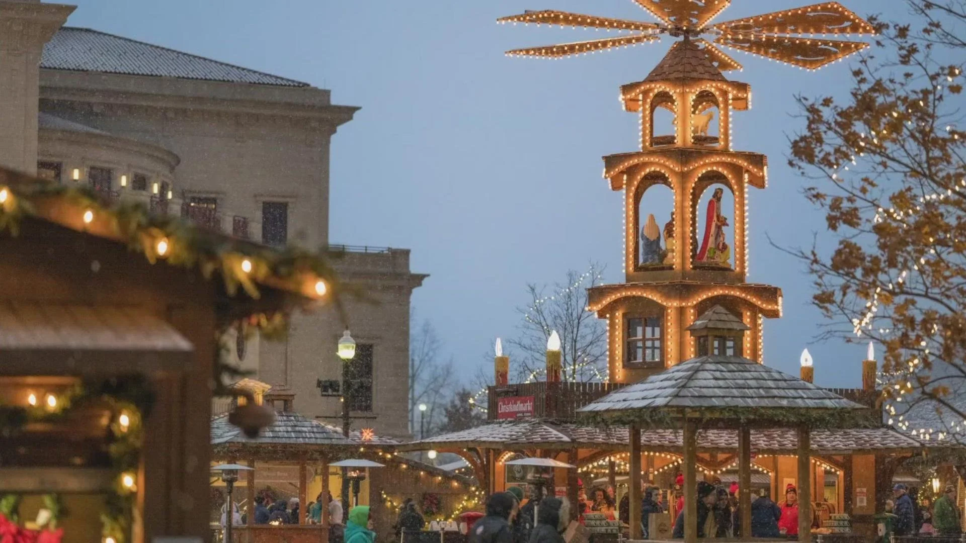 A festive Christmas market scene at dusk with a decorated tower displaying nativity figures, surrounded by wooden stalls with Christmas lights. People are browsing the market. Carmel Christkindlmarkt
