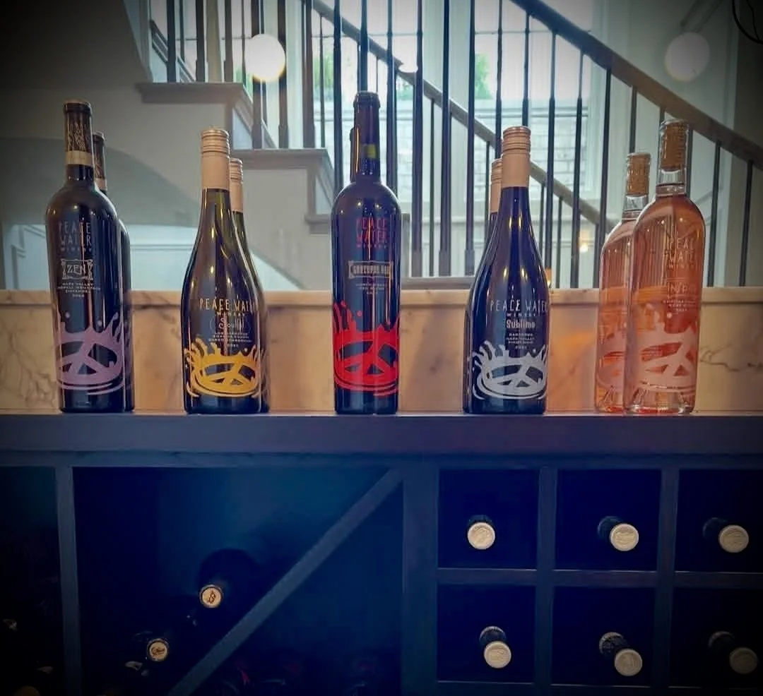 Five bottles of Peace Water wine in different colors arranged on a bar counter with a wine rack below, with a staircase and railing in the background. Wine tasting room. Carmel, Indiana. Napa wines.