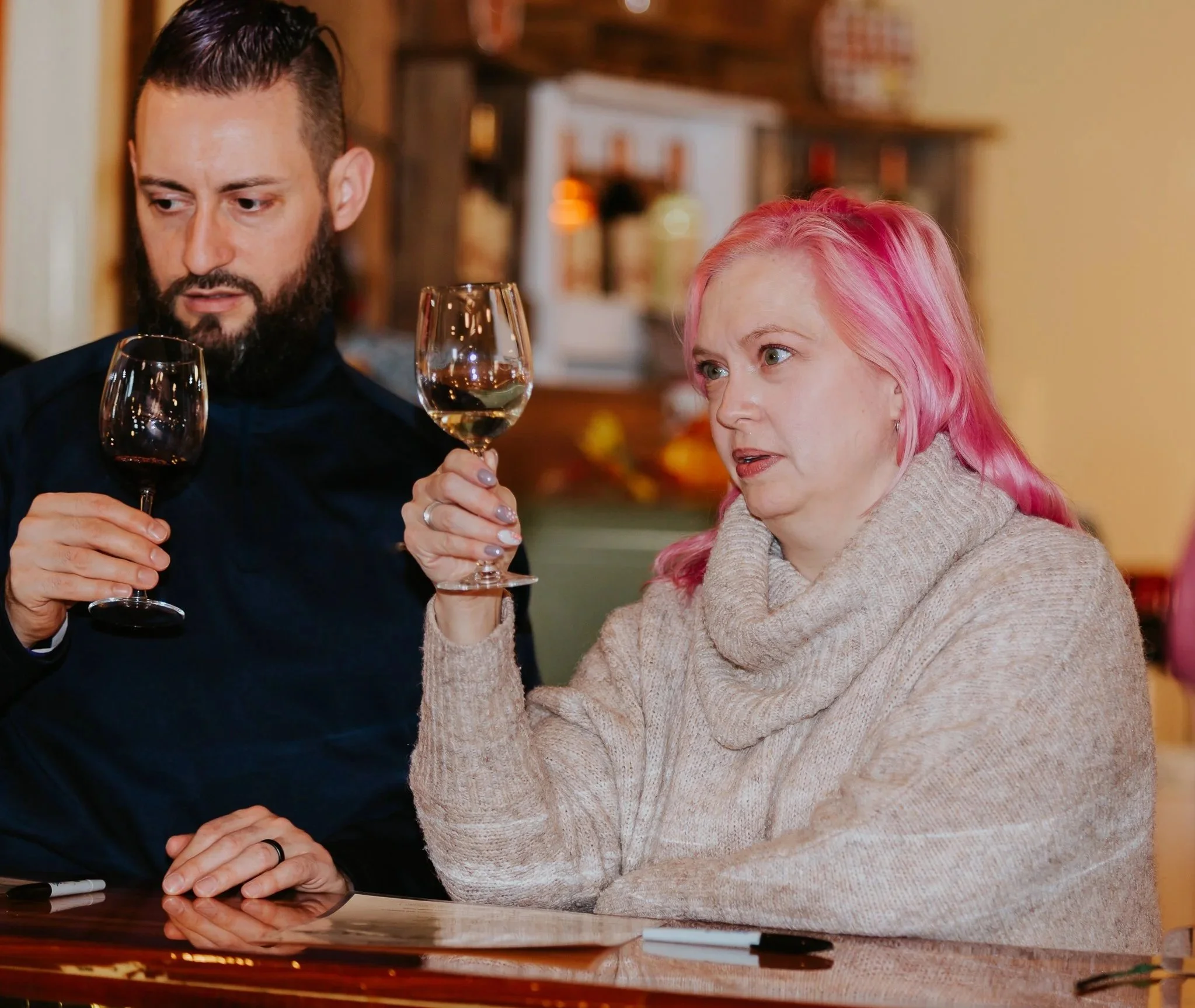 A man with a beard and a woman with pink hair sitting together, holding wine glasses, in a cozy indoor setting. Wine tasting experience. Wine sampling.