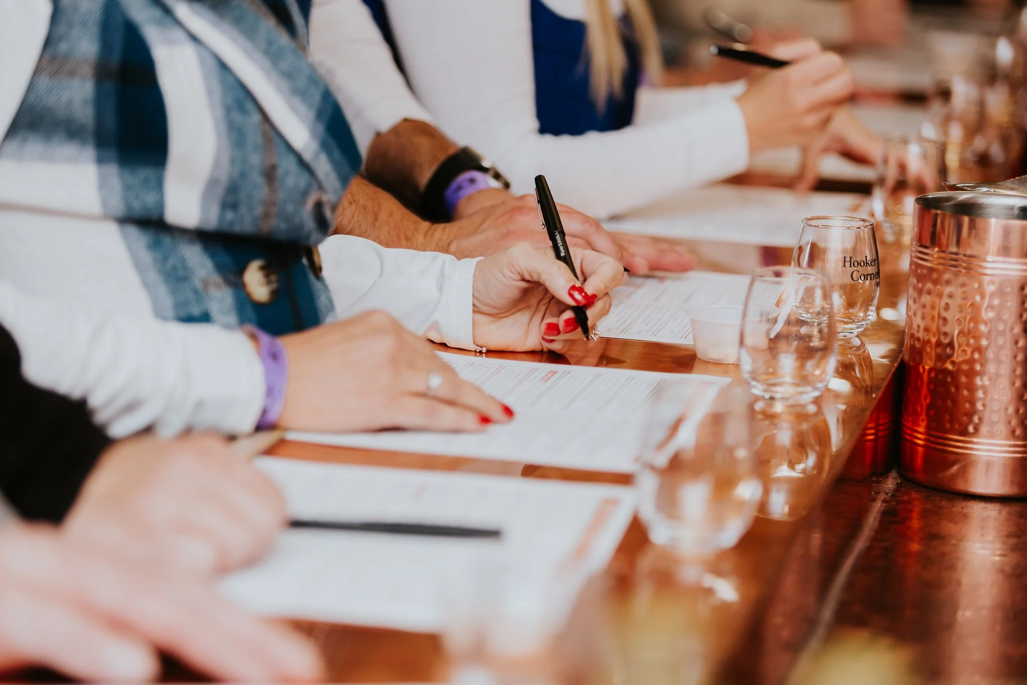 People at a wine tasting bar with wine sampling menus, wine tasting glasses, and a copper container, some taking notes or using their phones. Custom wine tasting experience. hand selected wines.