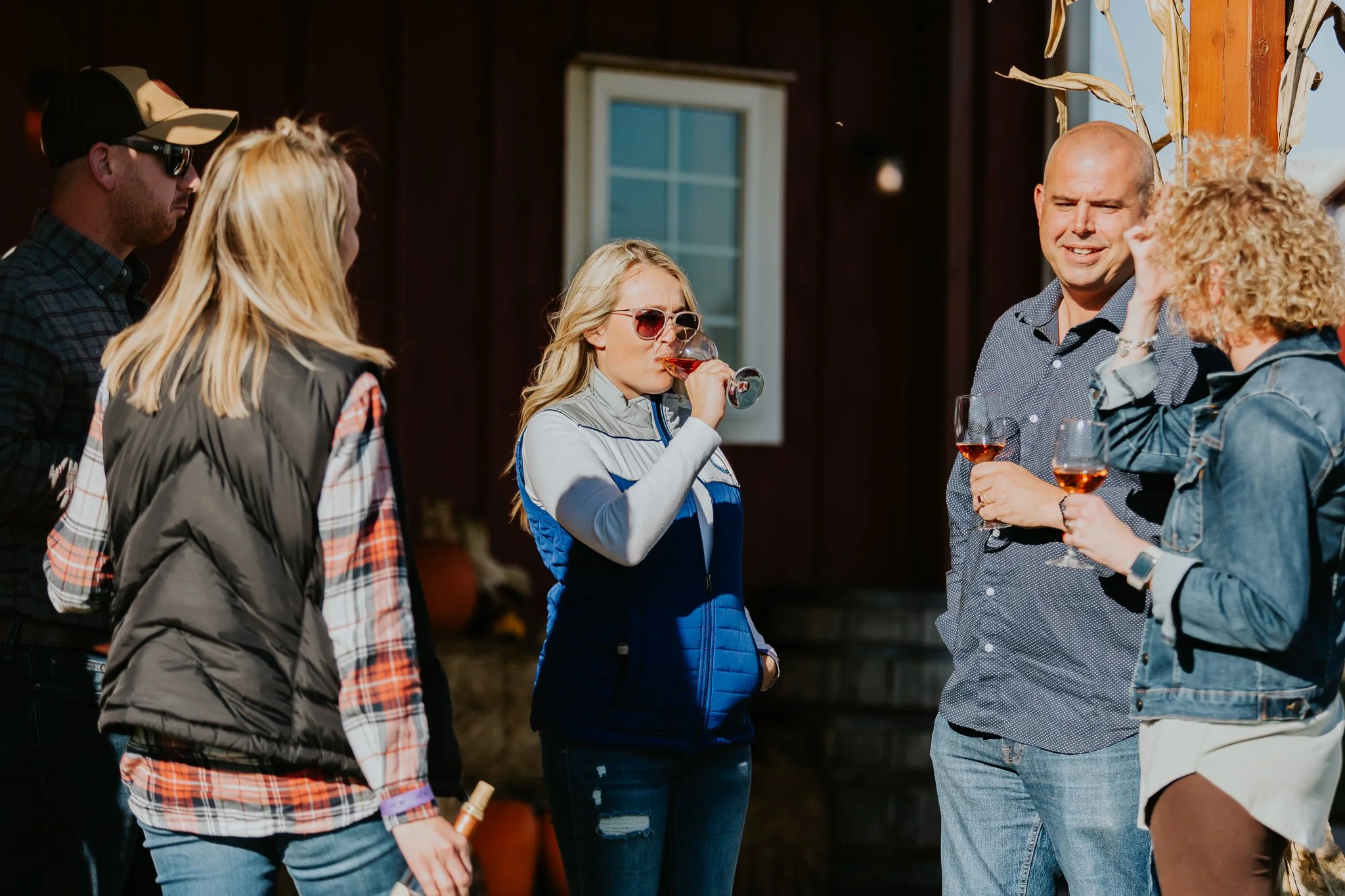 Group of people socializing outdoors, holding glasses of rosé wine, one woman sipping from her glass, wearing casual clothing with some wearing sunglasses and plaid shirts. Lovely autumn day enjoying wine with friends. Men and women drinking wine.