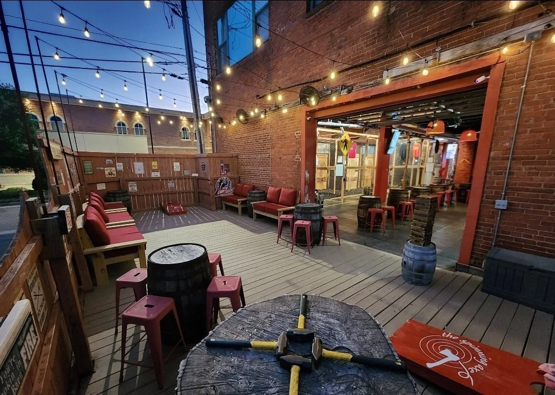 Outdoor patio with wooden decking, string lights, red cushioned benches, small tables with red metal stools, and a brick building with a bar area. Free to play outdoor patio games including corn hole, giant Jenga, ring toss, and Hammerschlagen.