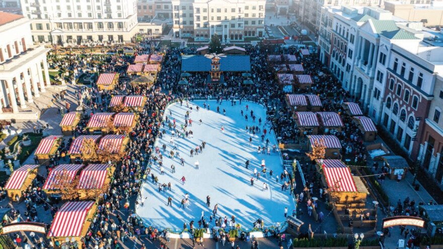 A bustling outdoor ice skating rink surrounded by market stalls with red and white striped roofs, people skating on the ice and walking around, in an urban shopping district with large buildings. Carmel Christkindmarkt