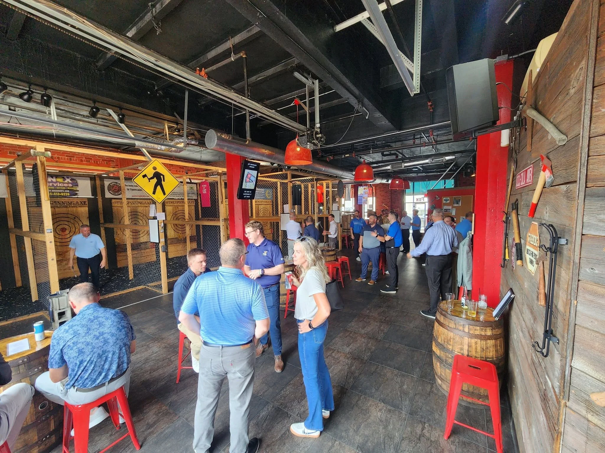 People standing and sitting inside axe throwing venue and bar with exposed brick walls, wooden decor, red pendant lights, and a sign for corn hole game on the wall.