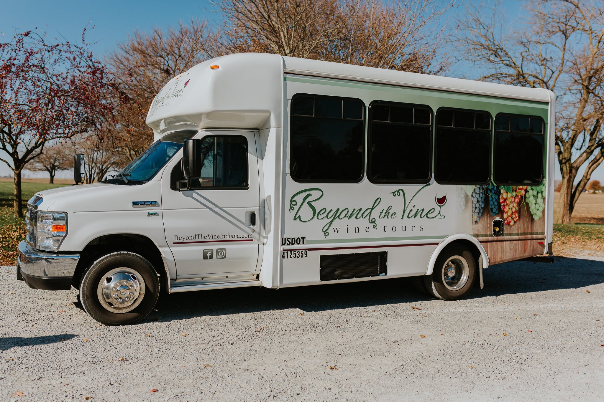A white shuttle bus with the logo 'Beyond the Vine' and grapevine artwork, parked outdoors with leafless trees and a clear blue sky in the background. Concert and special event transportation