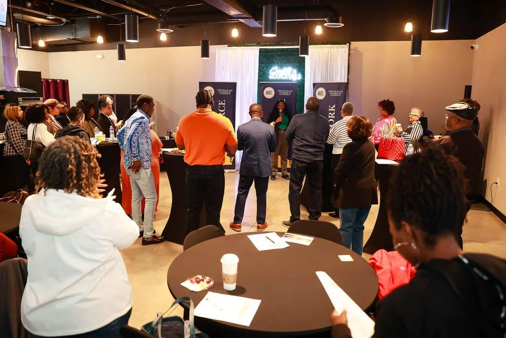 People attending a social or networking event in a conference room, with some standing and listening to a speaker at the front. There are tables and banners with the word 'SOCIAL' visible.