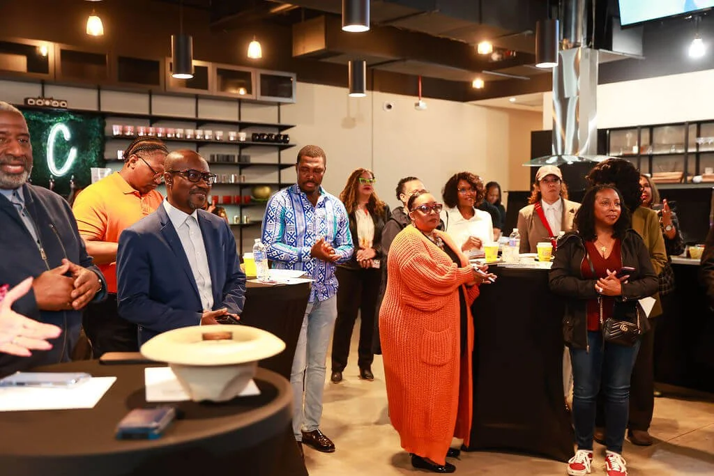 Group of diverse people gathered in a modern indoor space, some standing and listening attentively, with drinks on the counter and a blackboard in the background.