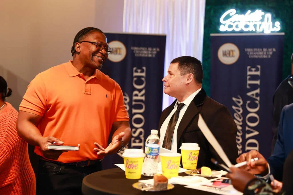 Two men engaged in conversation at a networking event, with a table holding coffee cups, a water bottle, and snacks, and a backdrop with signs related to resources and advocacy.