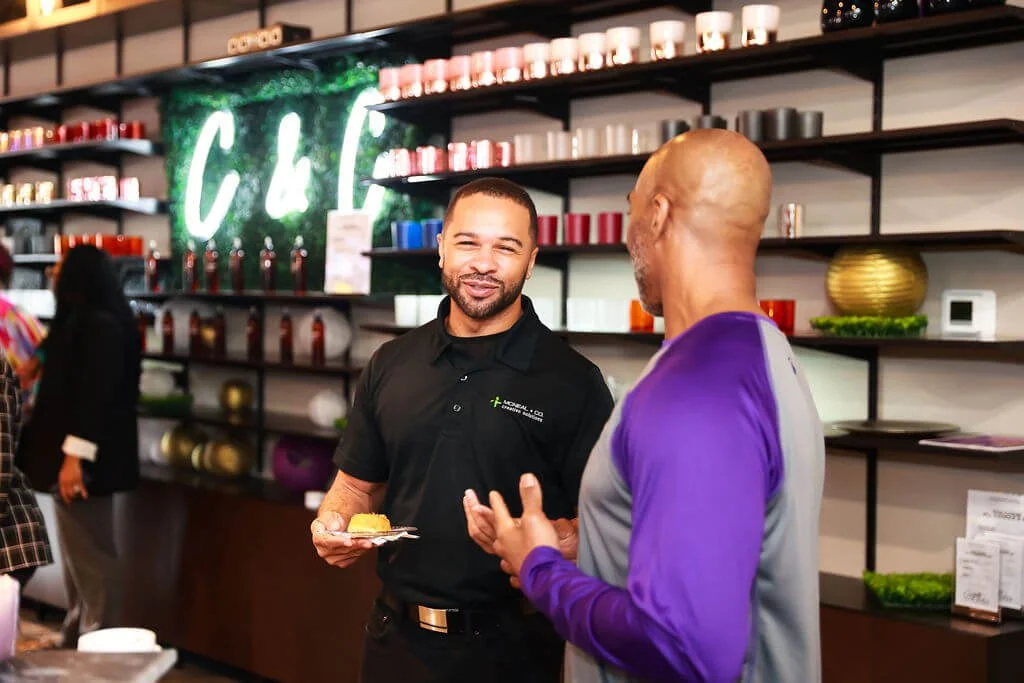 Two men are having a conversation in a retail store with shelves displaying various colored items behind them. One man, holding a plate with a snack, is smiling. The other man, wearing a purple and gray shirt, is listening. There are more people and 