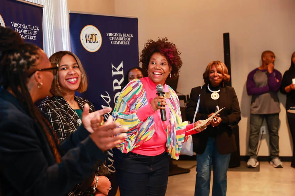 A woman with curly hair holding a microphone and a paper, smiling at a gathering organized by the Virginia Black Chamber of Commerce, with several women and a few men standing nearby.