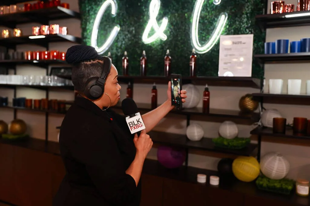 A woman with headphones and hoop earrings is holding a microphone and a smartphone, recording or taking a picture in front of a decorated wall with a neon sign that says 'C & C' and various decorative bottles and objects on shelves.