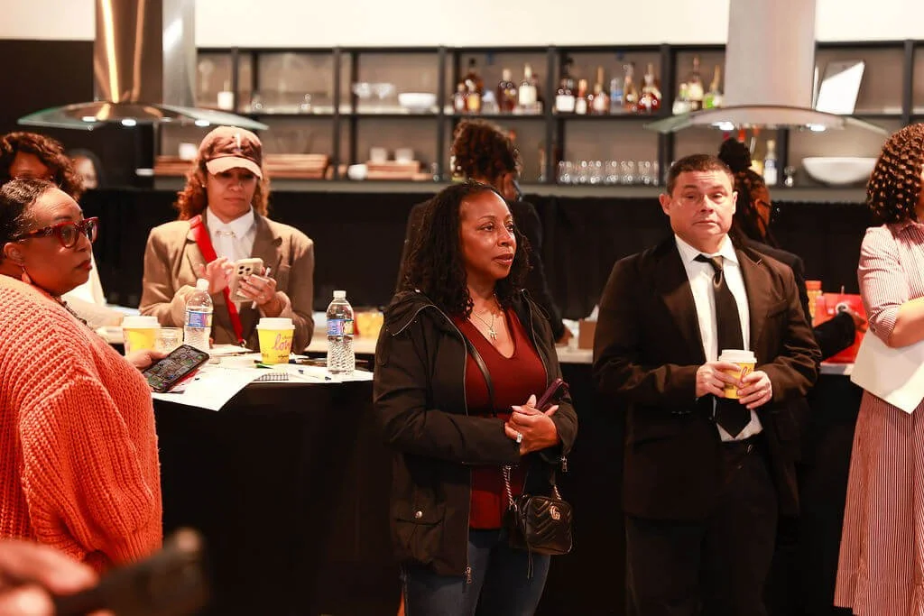 People attending a social event or gathering, standing near a bar or counter, holding drinks, with shelves of bottles and glasses in the background.