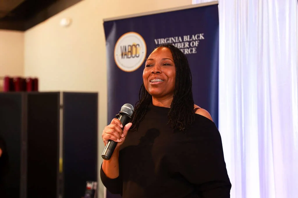 A woman smiling and speaking into a microphone at an indoor event with a Virginia Black Chamber of Commerce banner in the background.