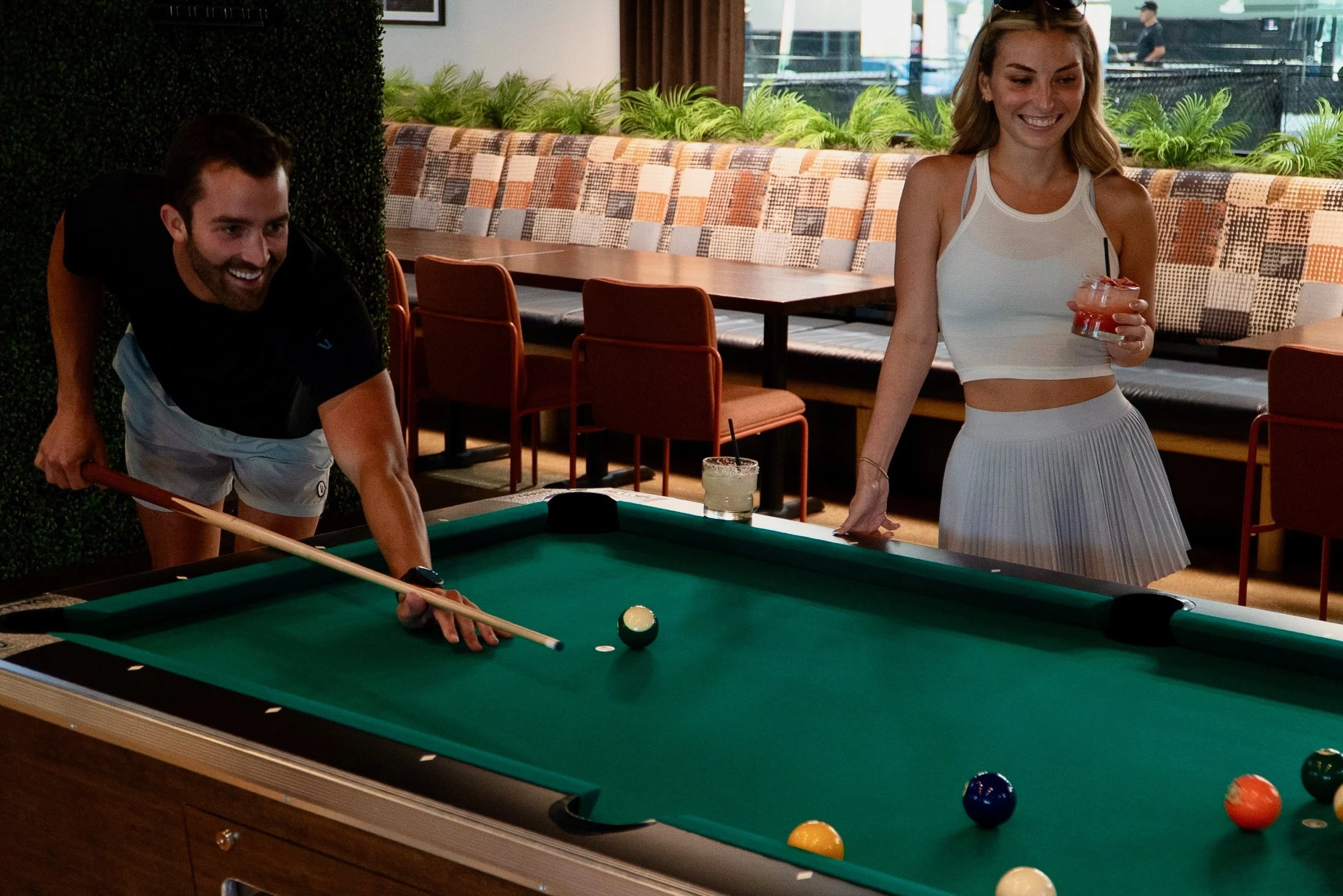 A man and a woman playing pool at a bar or restaurant, with the man aiming a cue stick at the cue ball and the woman smiling while holding a drink.
