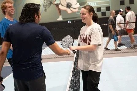 Two women at an indoor pickleball court, one handing a paddle to the other, with a man laughing nearby and other people playing in the background.