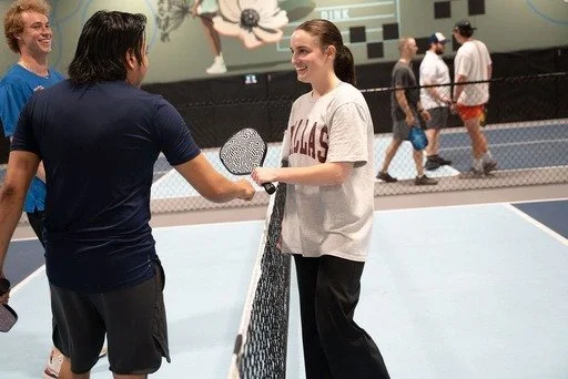 Two women shake hands on a tennis court, smiling. Other people in the background are walking or standing.