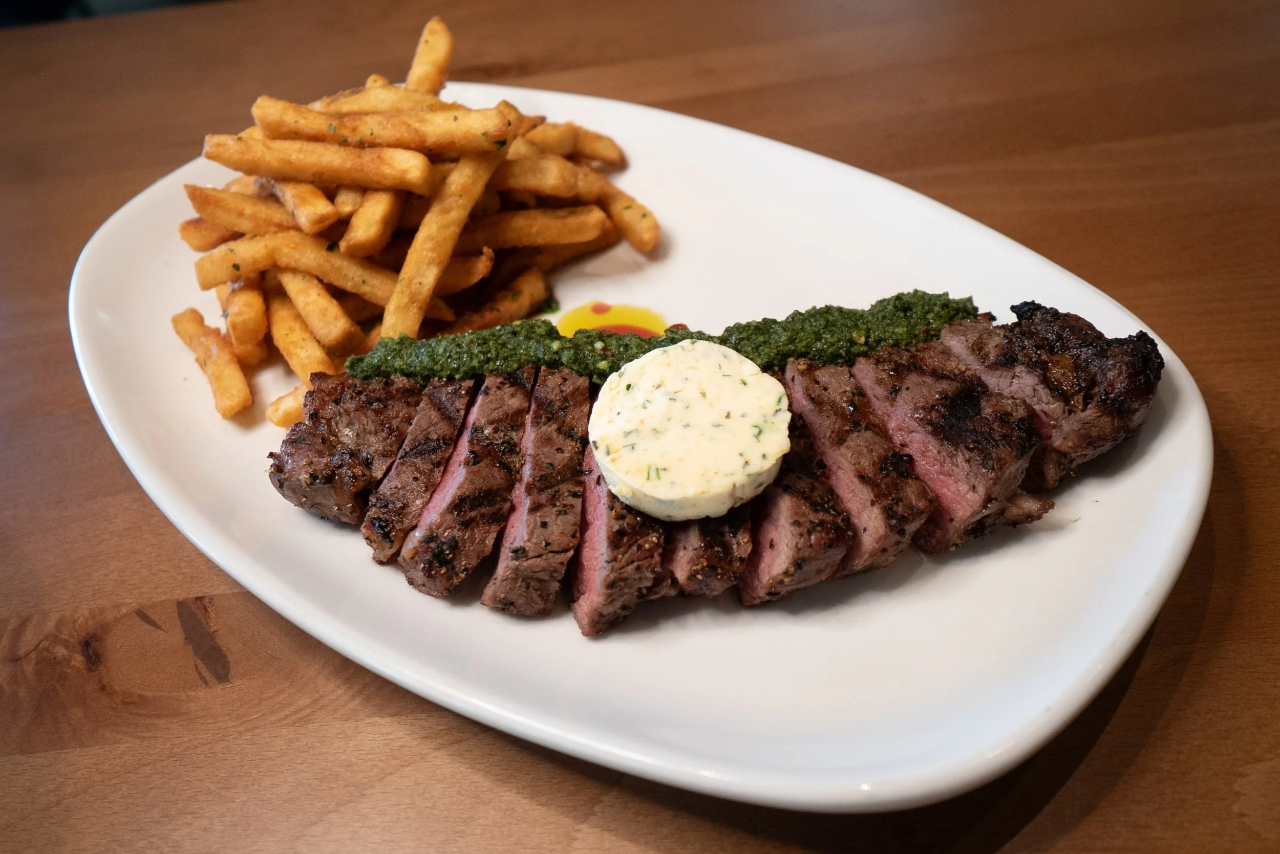 A plate with sliced grilled steak topped with herb butter, green chimichurri sauce, beside a side of French fries. The dish is garnished with green sauce and a small drop of yellow sauce.